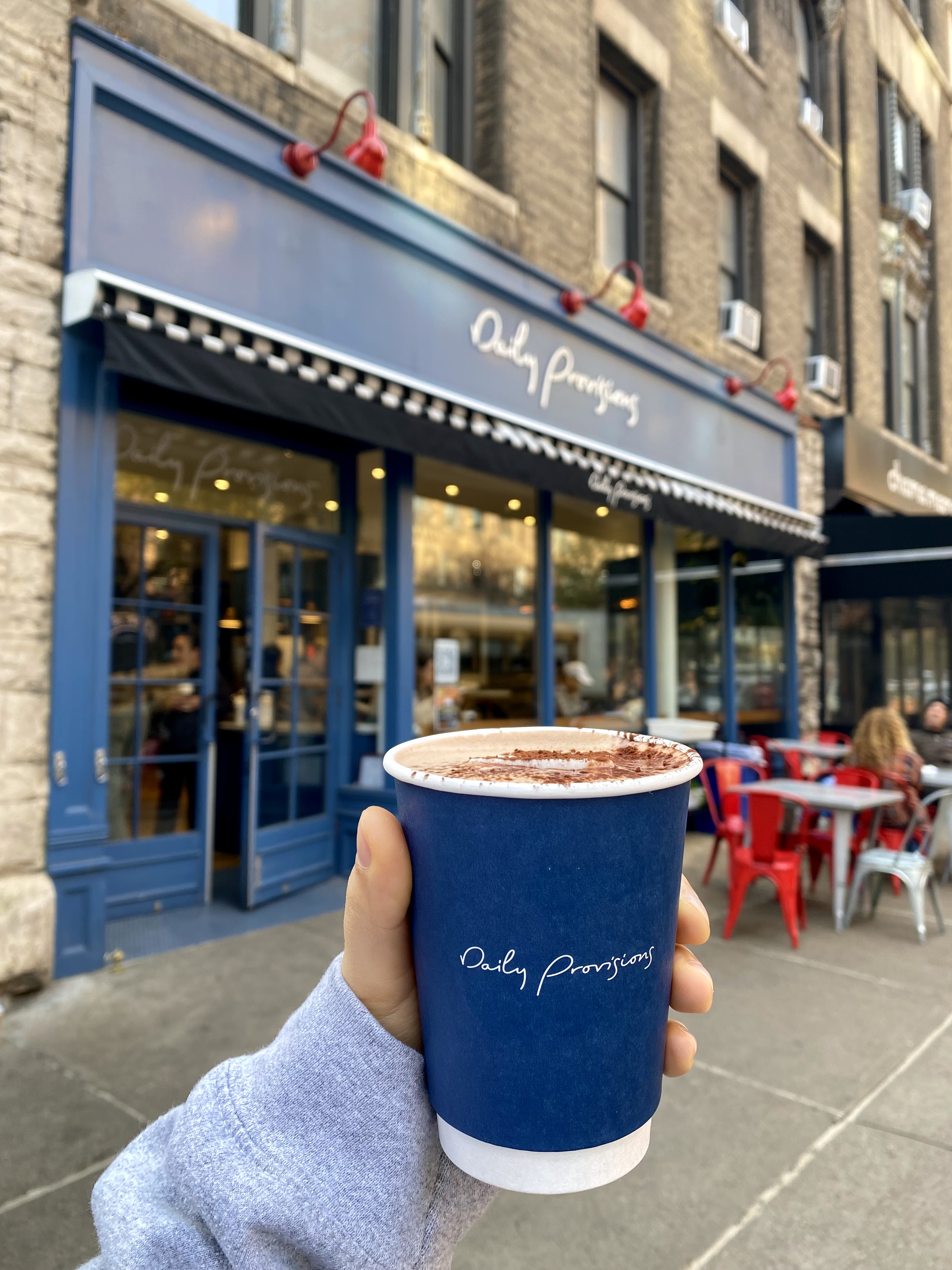 A hand holds a cup of hot chocolate outside the Daily Provisions storefront.