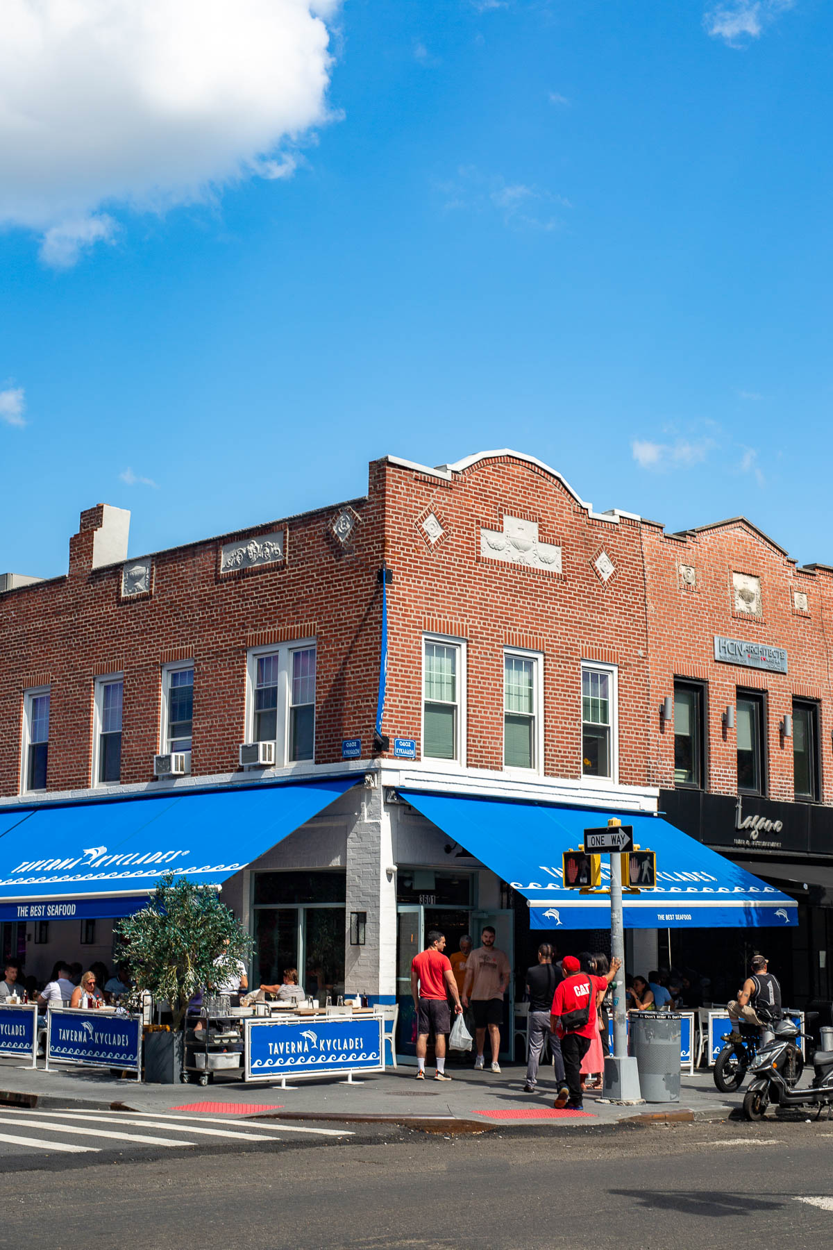 A brick corner building with blue awnings houses Taverna Kyclades restaurant on an Astoria street corner.