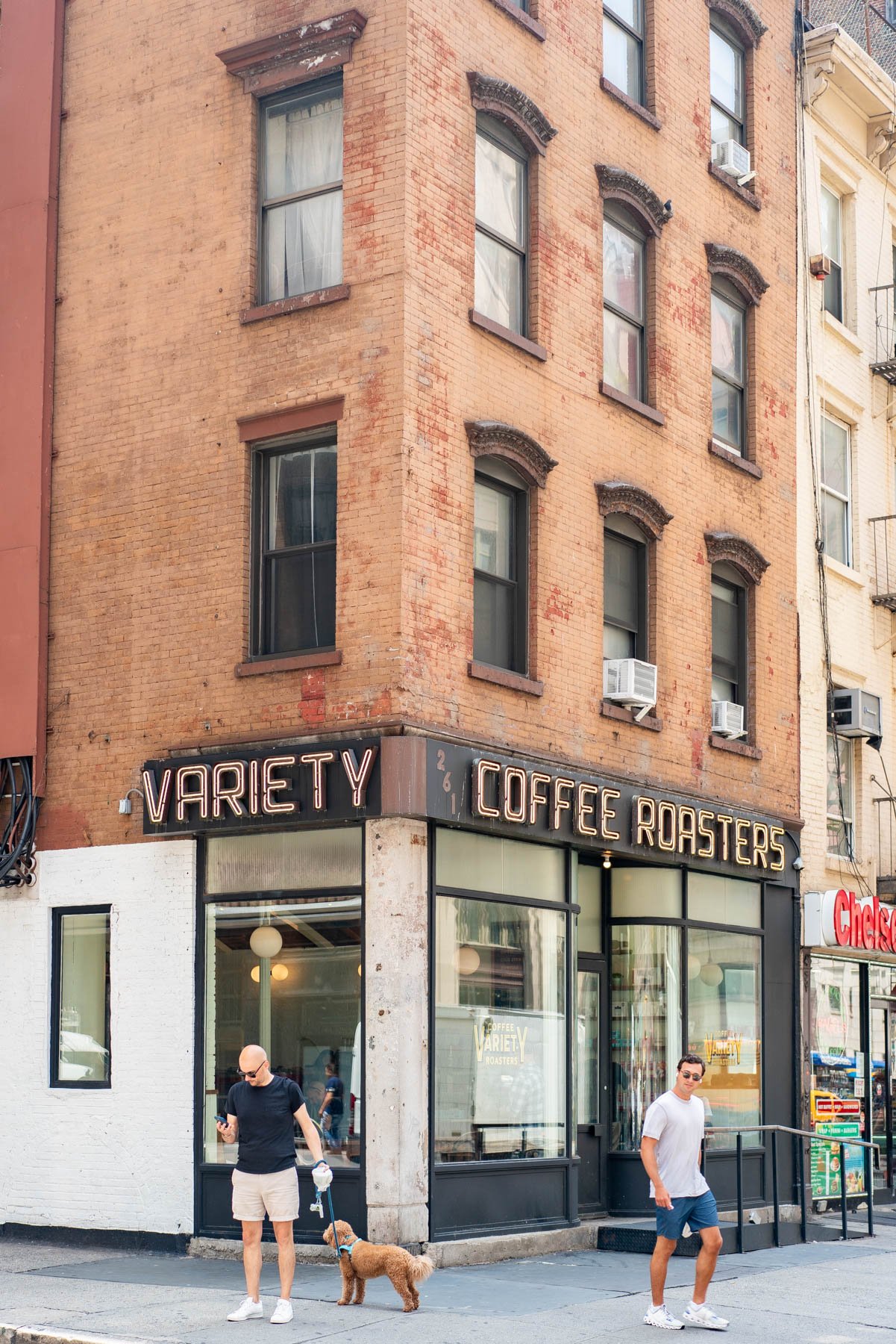 A brick corner building houses Variety Coffee Roasters with people and a dog walking past on the sidewalk.