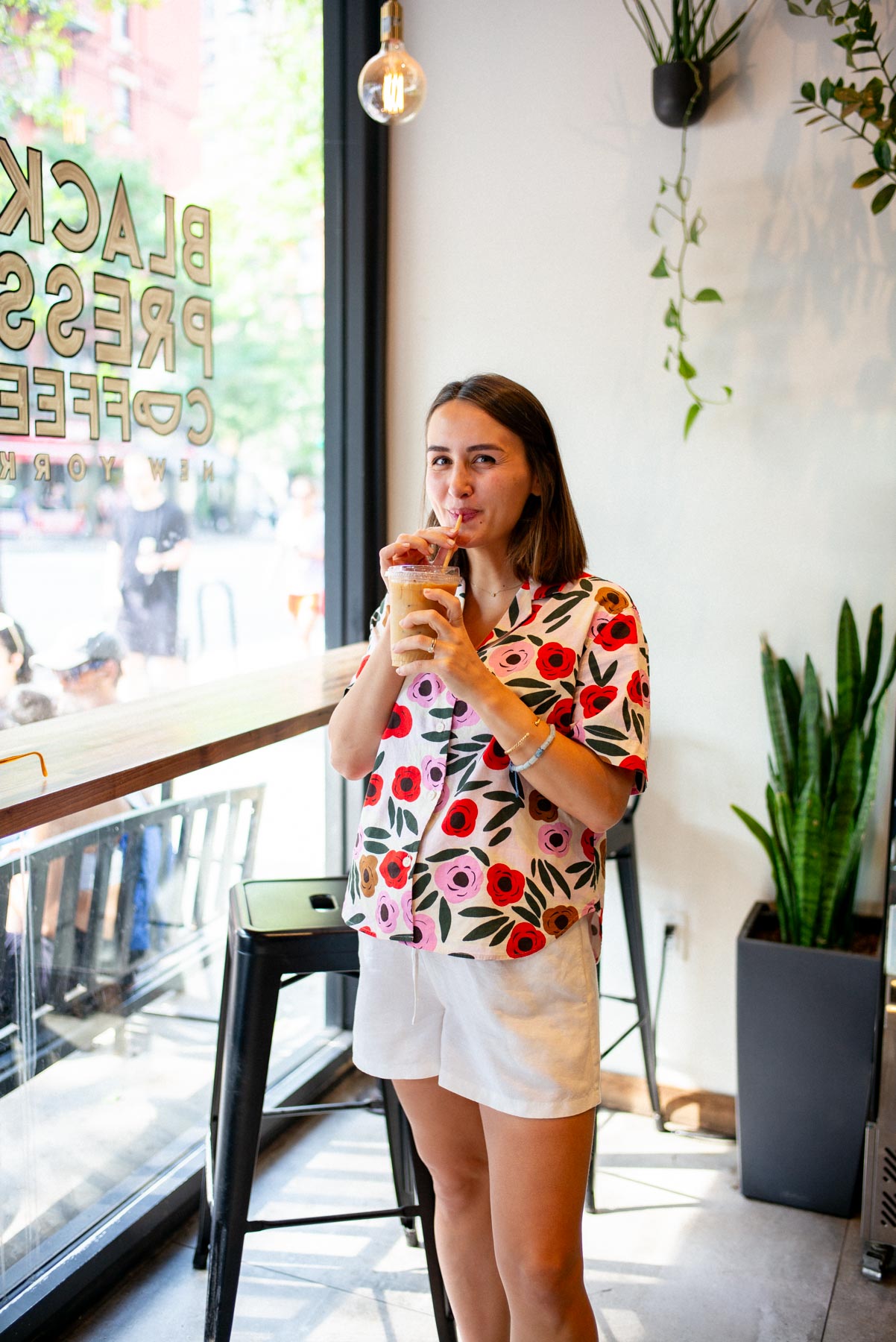 A woman drinks an iced coffee inside Black Press Coffee near the window.