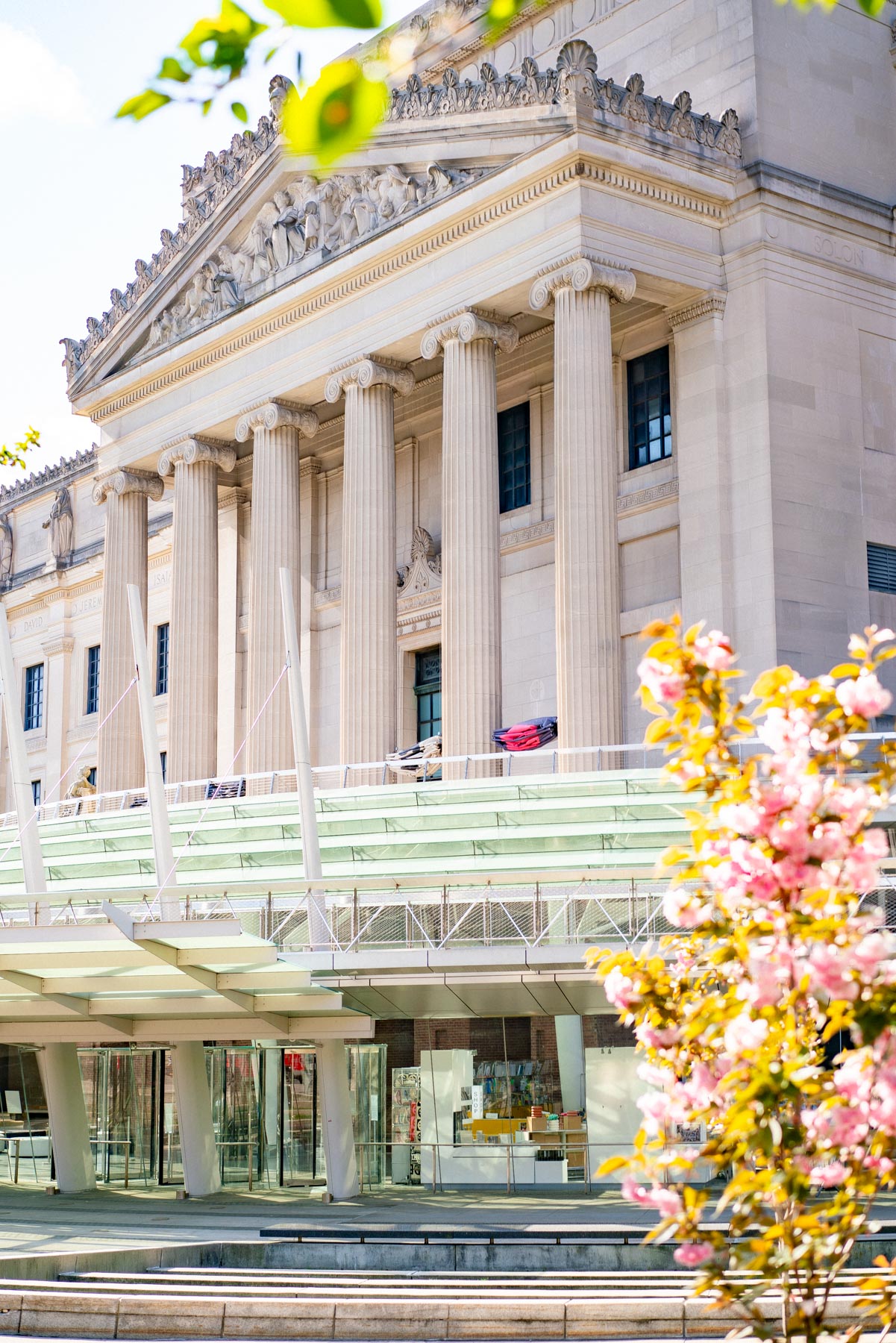 Exterior shot of the Brooklyn Museum is made with intricately carved architecture and large stone pillars surrounded by spring blooms and glass windows.