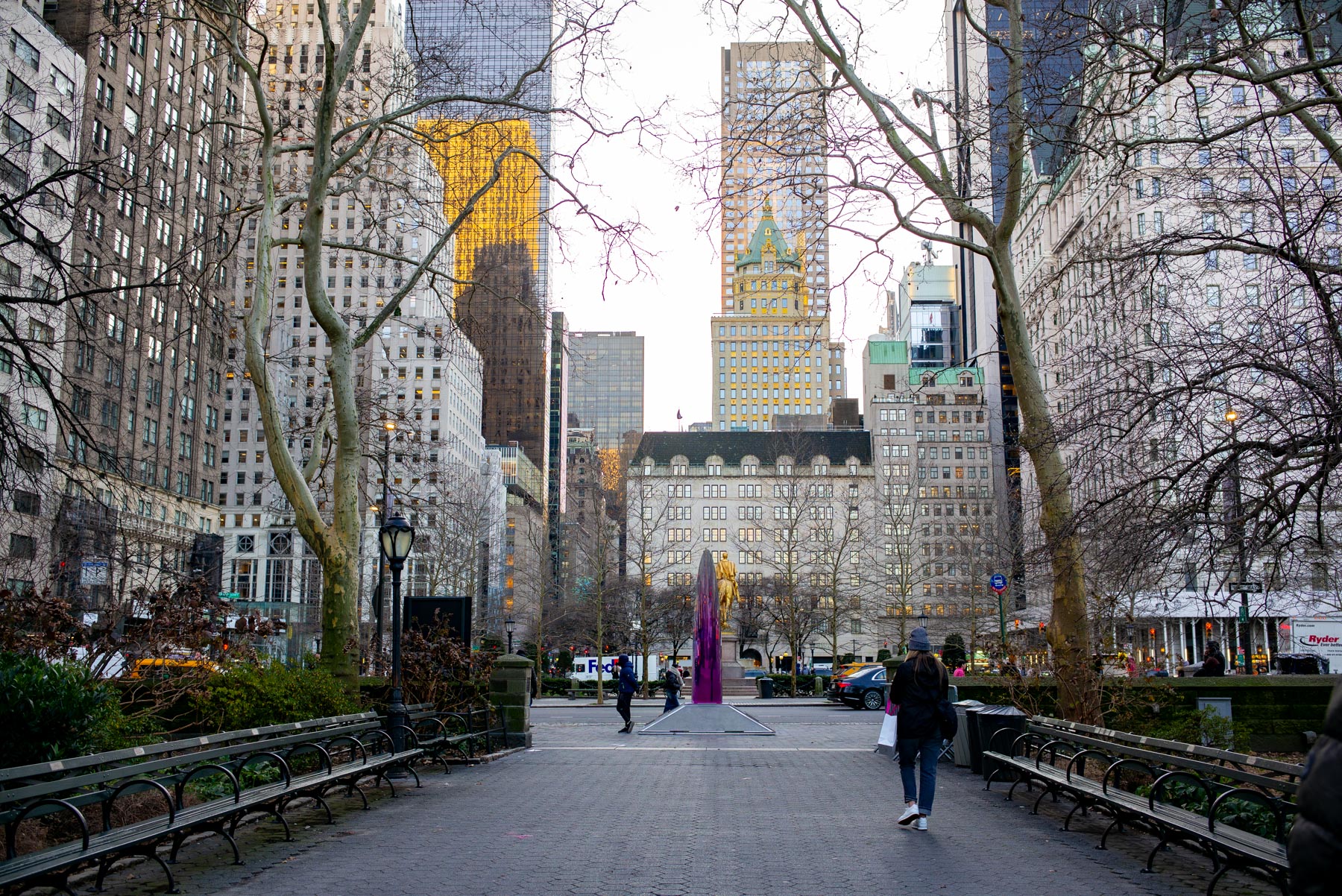 A view down a tree-lined park promenade toward Fifth Avenue skyscrapers with a gold-topped tower in the distance.