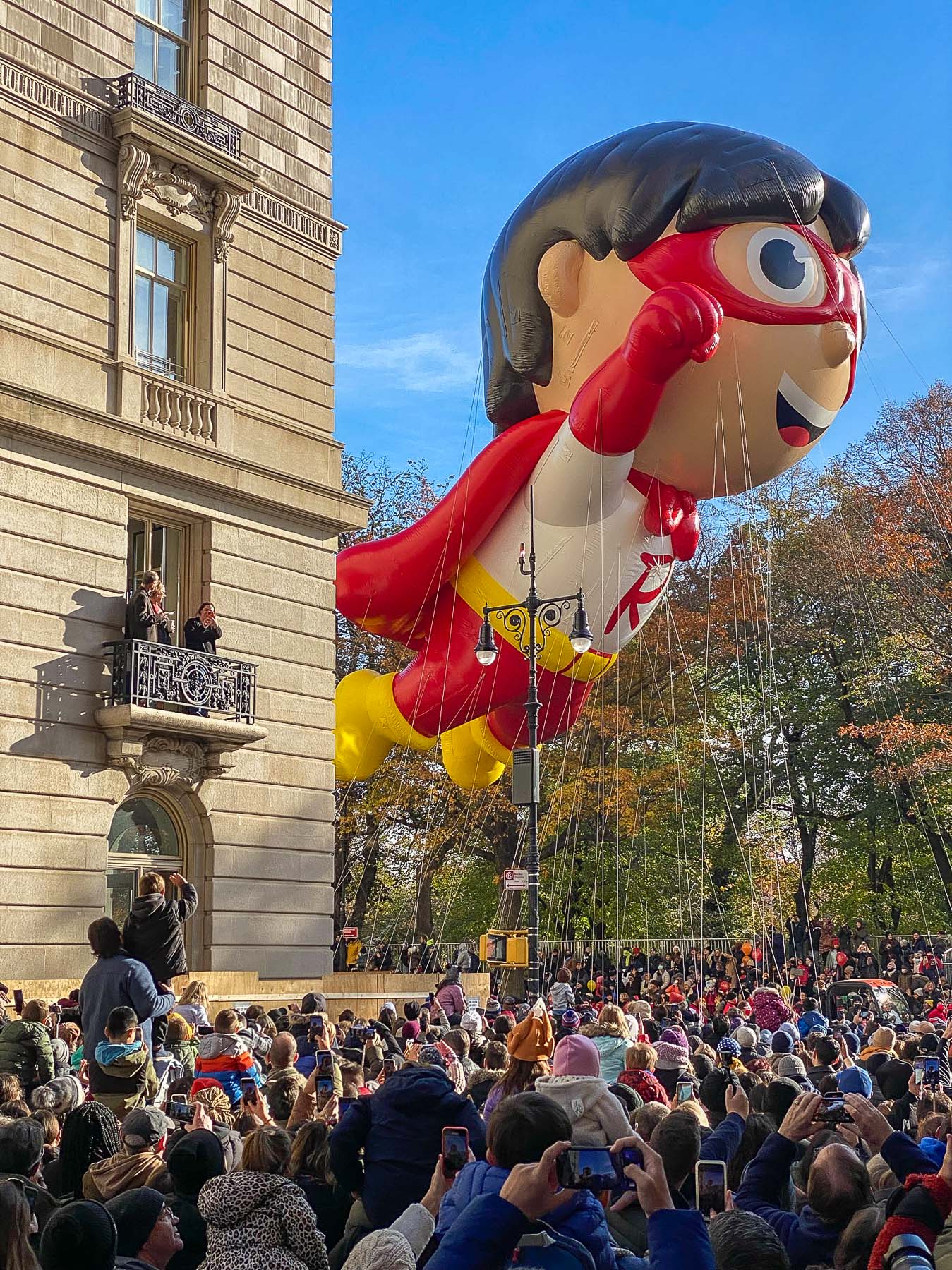 A giant superhero character balloon floats past an apartment building during the Macy's Thanksgiving Day Parade as crowds watch from the street and a balcony.
