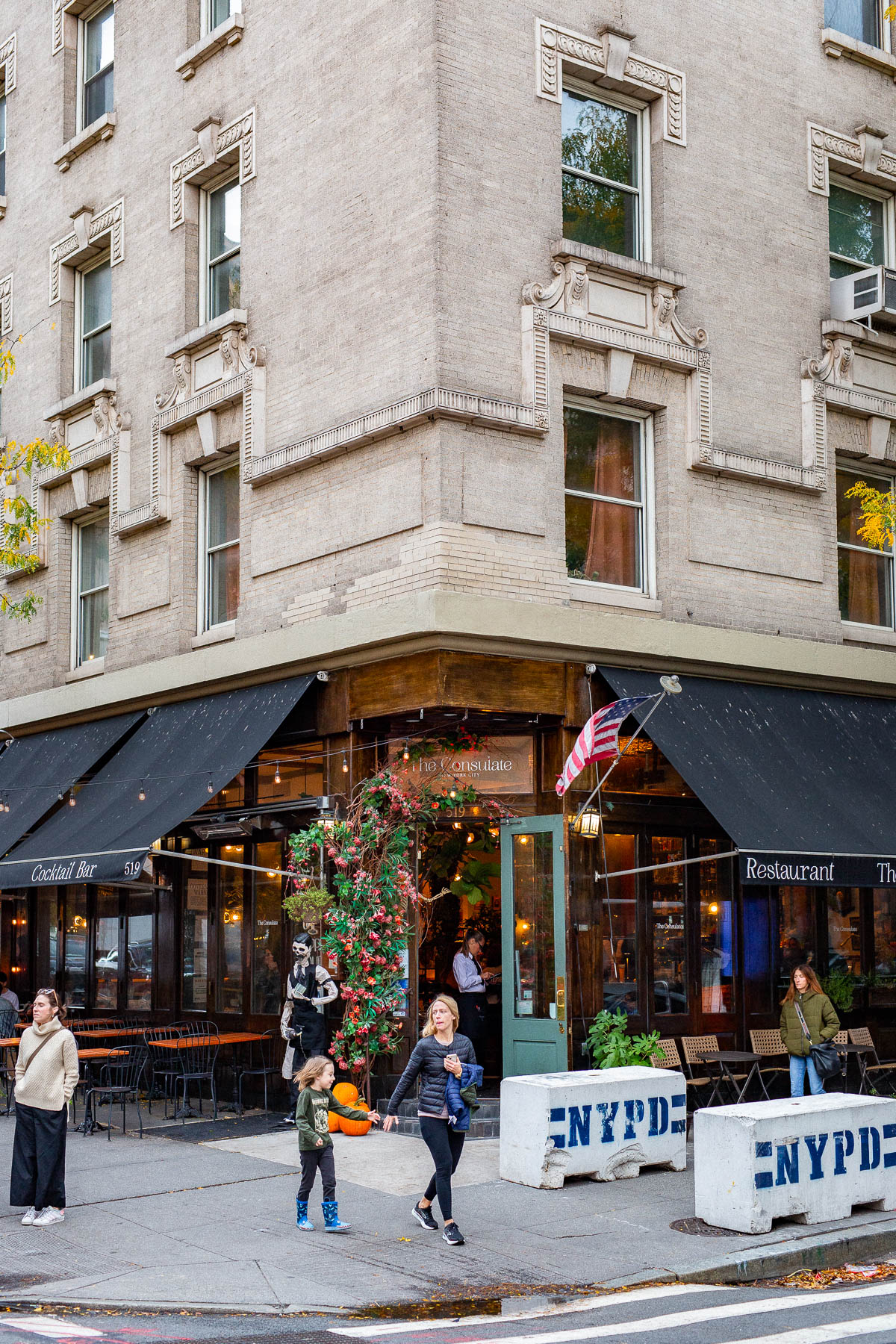 A corner restaurant called The Consulate sits beneath a stone building with people walking past on the sidewalk.