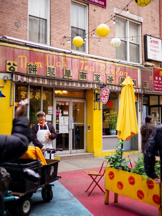 The storefront of Nom Wah Tea Parlor in Chinatown with a worker standing outside and paper lanterns strung overhead.