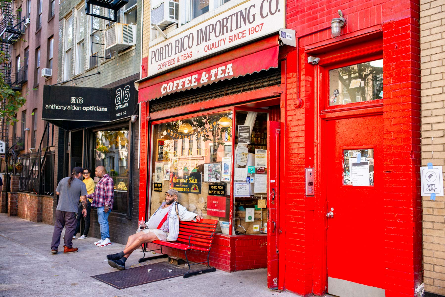 A man sits on a bench outside the Porto Rico Importing Co. coffee and tea storefront while others chat nearby.