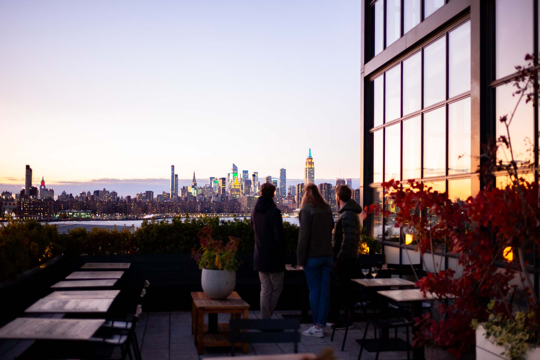 Three people stand on a rooftop bar watching the Manhattan skyline across the East River at dusk.