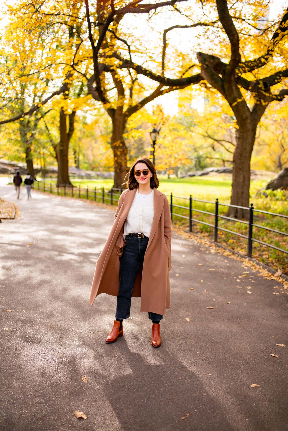 A woman in a camel coat stands on a park path lined with yellow autumn trees.