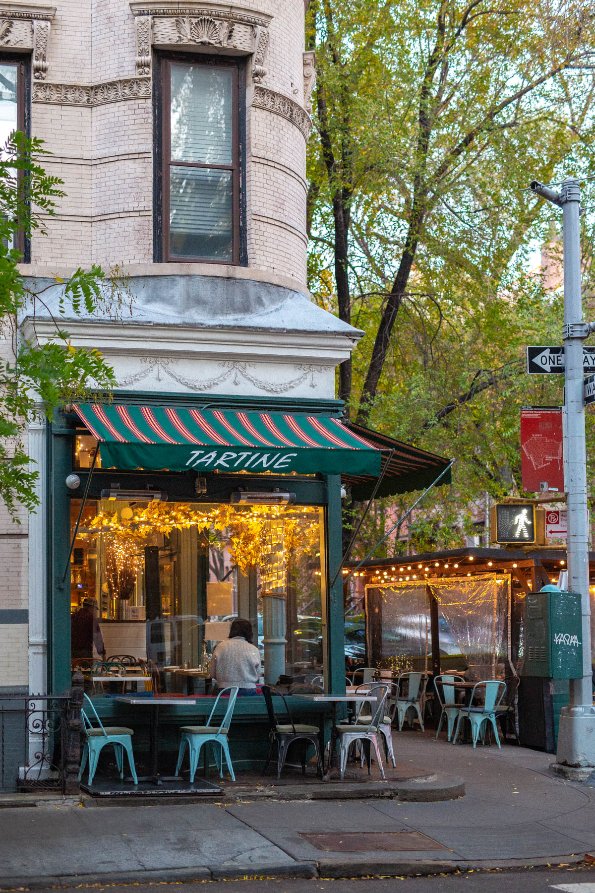 A corner restaurant with a striped awning and outdoor tables sits along a tree-lined West Village street.