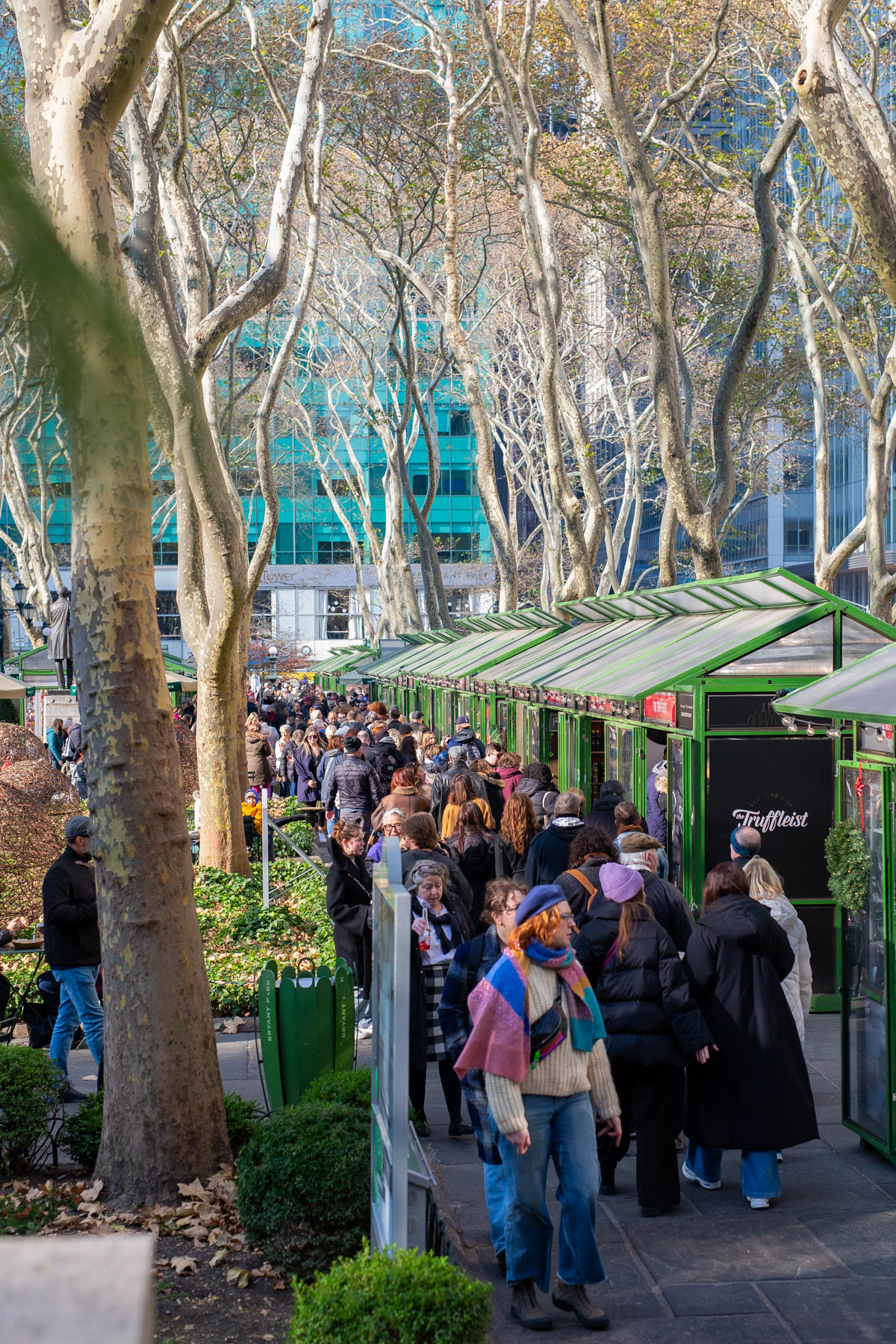 Crowds walk past green market stalls under bare trees at Bryant Park Winter Village.
