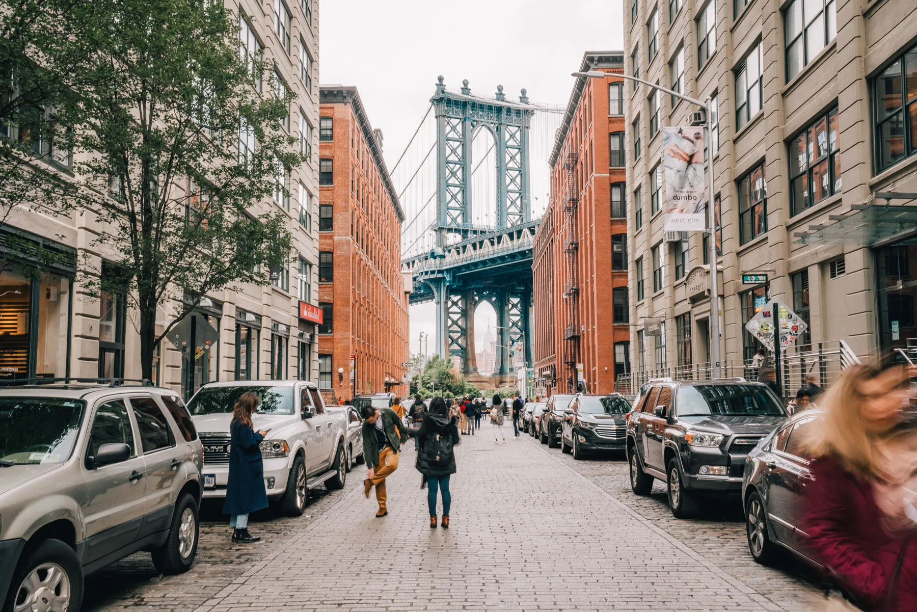 Pedestrians walk down a cobblestone street in Dumbo with the Manhattan Bridge framed between buildings.