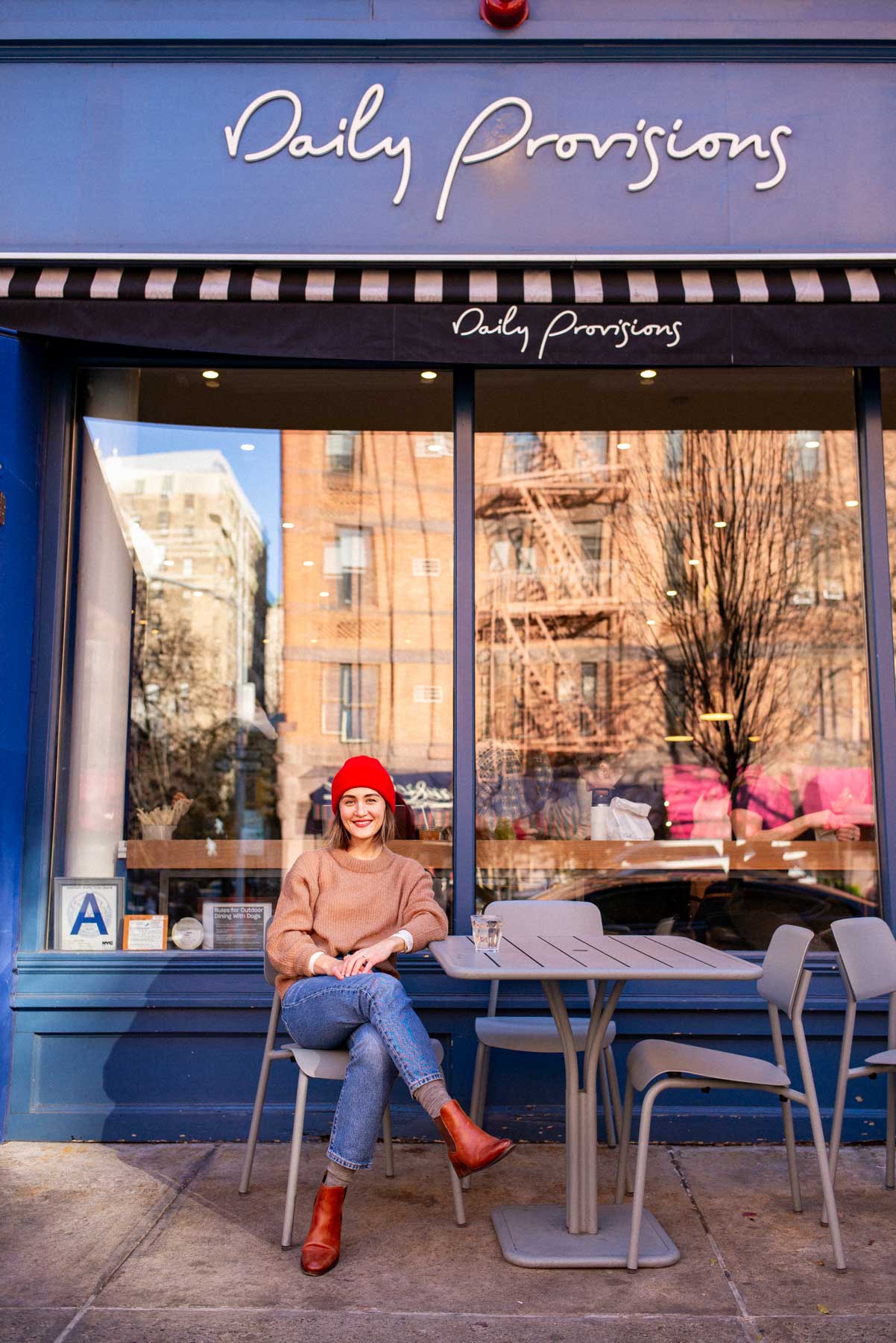 A woman in a red beanie sits at an outdoor table in front of the Daily Provisions storefront.