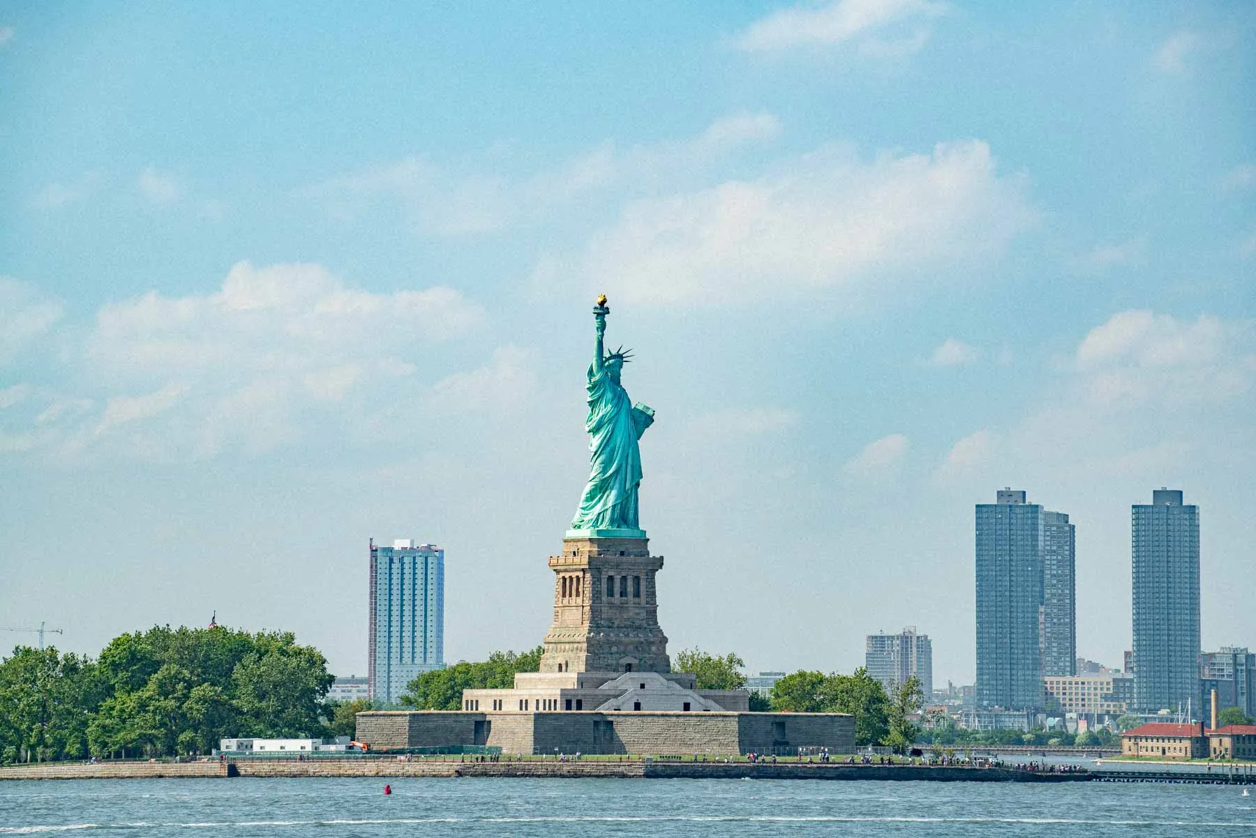 The Statue of Liberty stands on its island with the harbor and skyline in the background.
