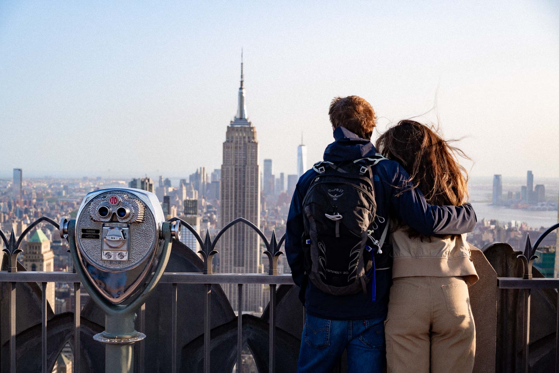 A couple stands at an observation deck railing looking out at the Empire State Building and Manhattan skyline.