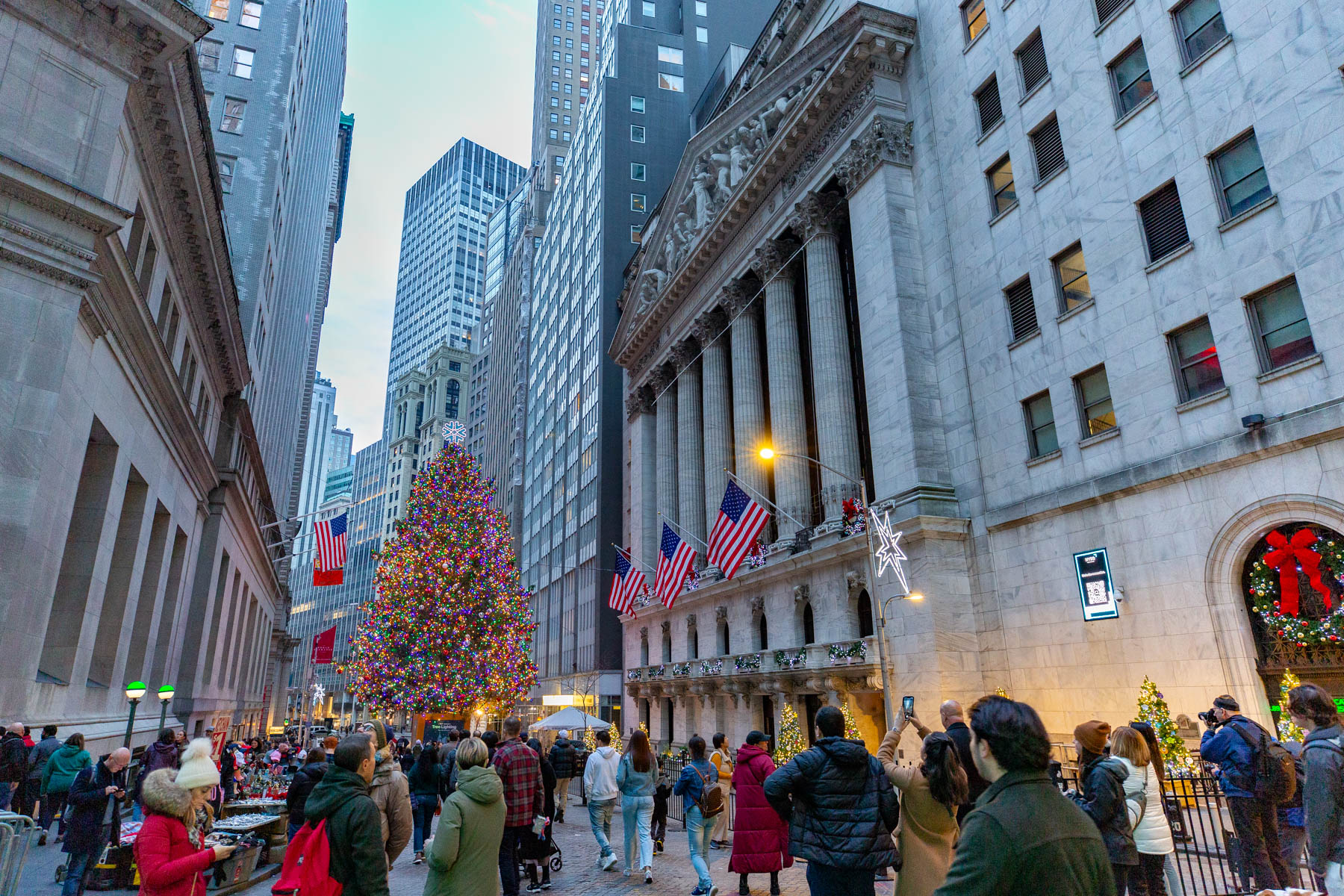 A large lit Christmas tree stands between the New York Stock Exchange and surrounding buildings on Wall Street as crowds gather.
