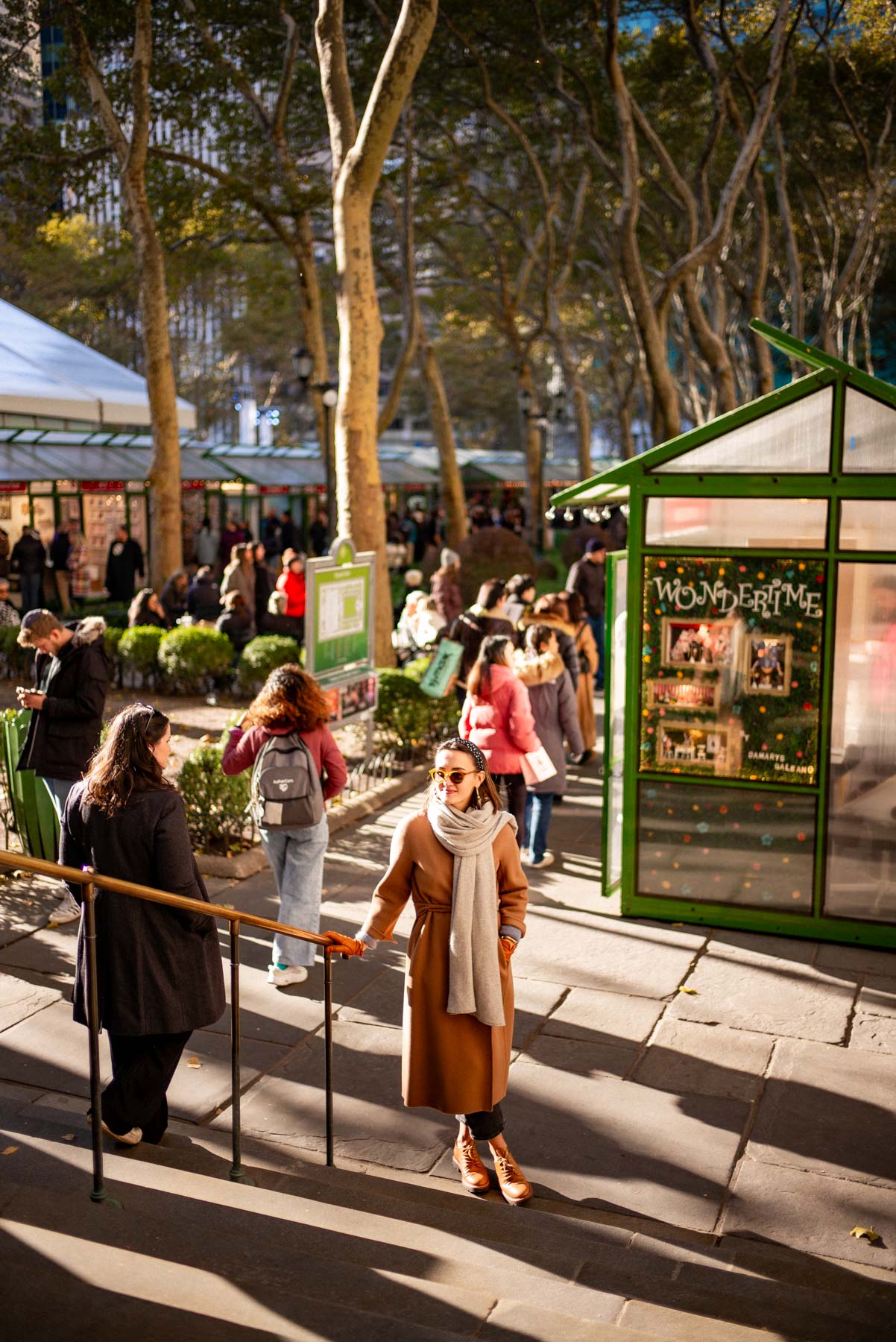 A woman in a camel coat walks through the holiday market at Bryant Park amid green market stalls and crowds.