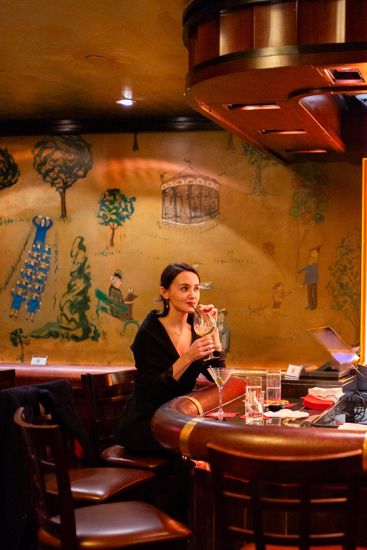 A woman sips a cocktail at the bar beneath the painted mural walls of Bemelmans Bar
