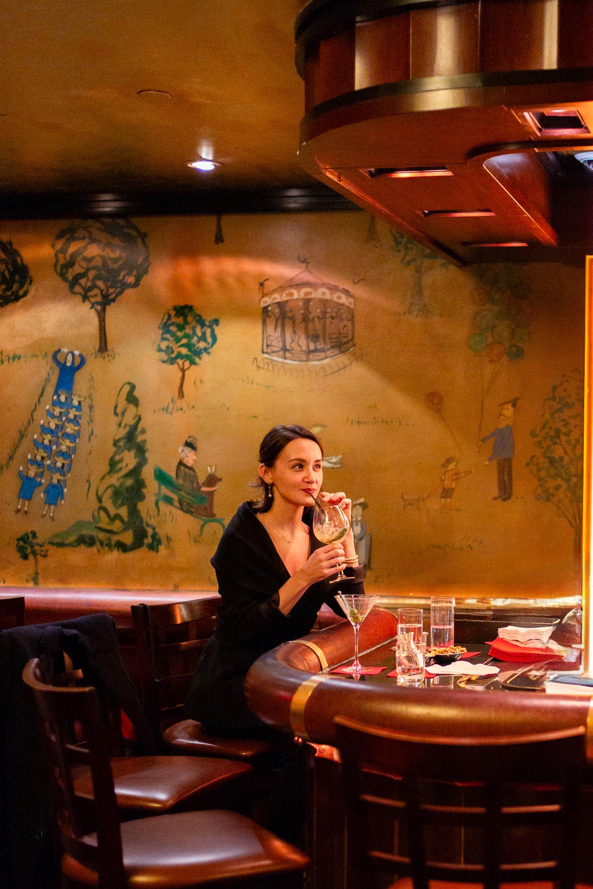 A woman sits at the bar sipping a drink beneath whimsical Central Park murals at Bemelmans Bar.