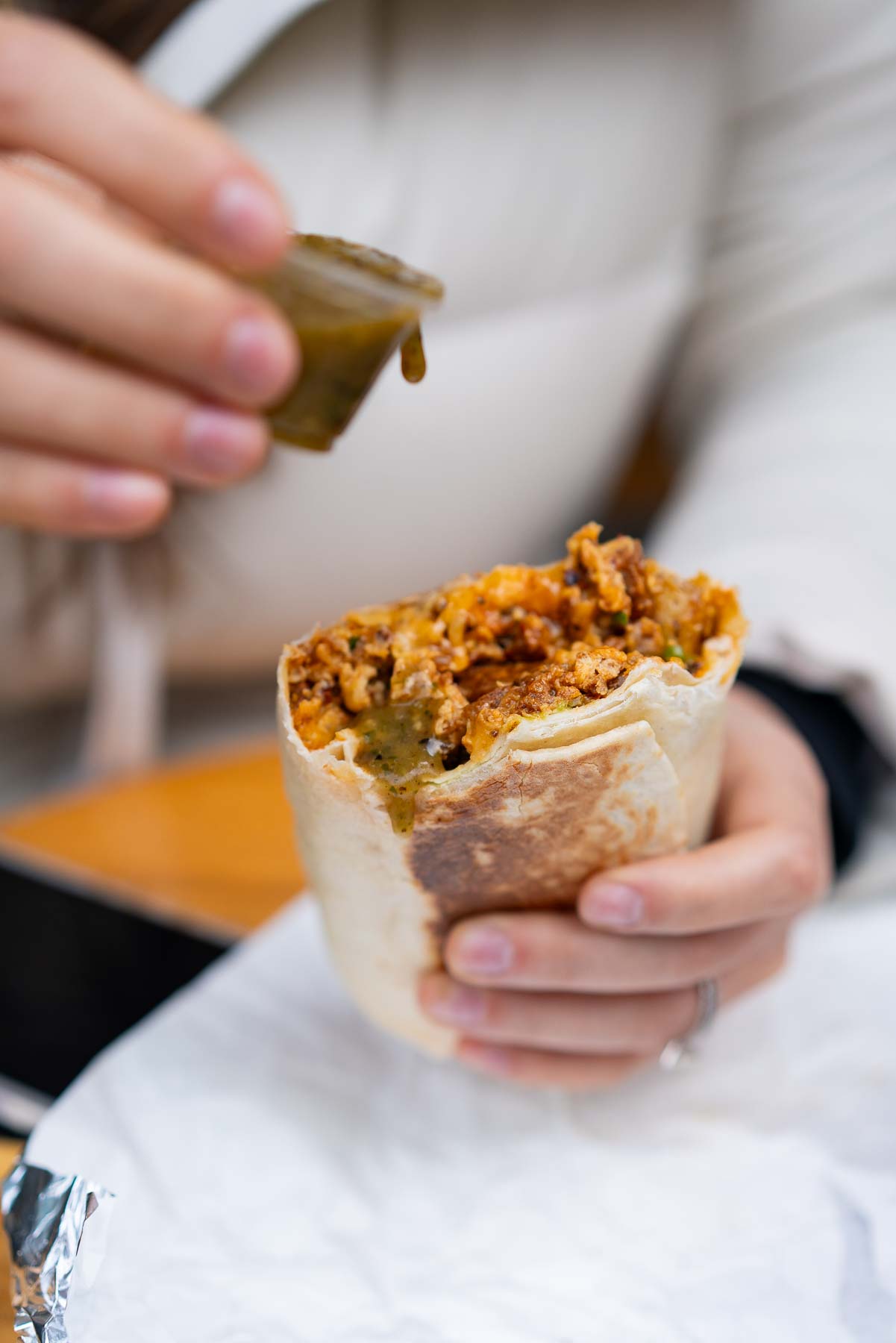 A person holds a breakfast burrito while squeezing green salsa from a small container onto it.