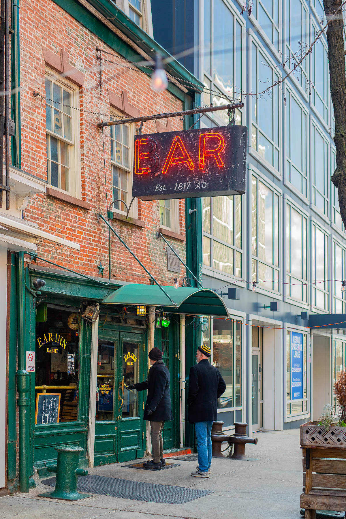 Two people stand outside the Ear Inn beneath its neon sign on a sunny day.