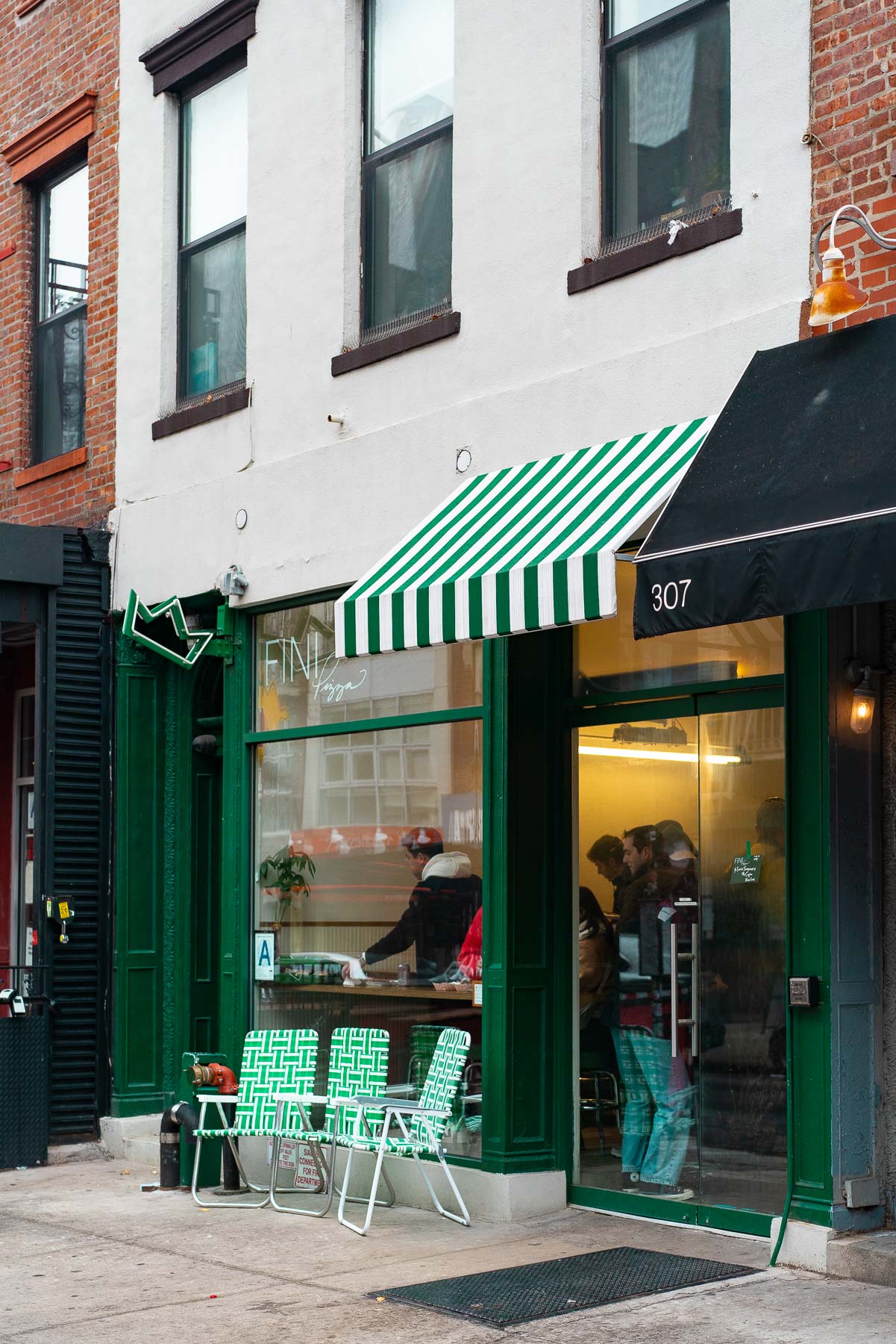 A pizza shop storefront with green striped awning and neon sign on a Williamsburg street corner.