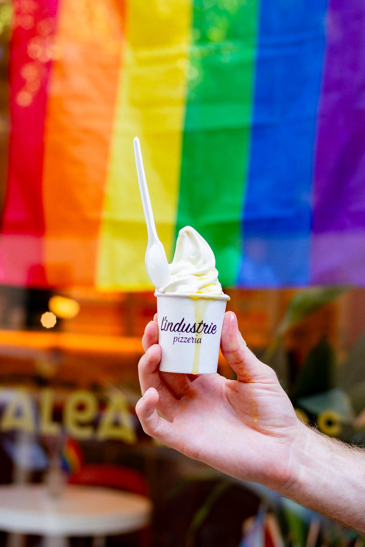 A hand holds a small cup of soft serve ice cream branded L'Industrie Pizzeria in front of a rainbow flag.