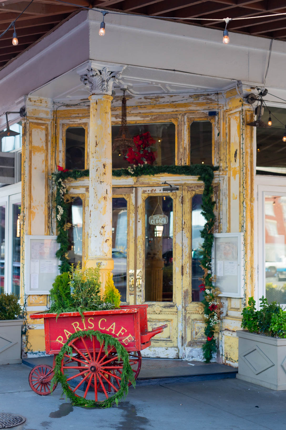 A weathered yellow storefront with a red cart reading Paris Cafe decorated with holiday garland.