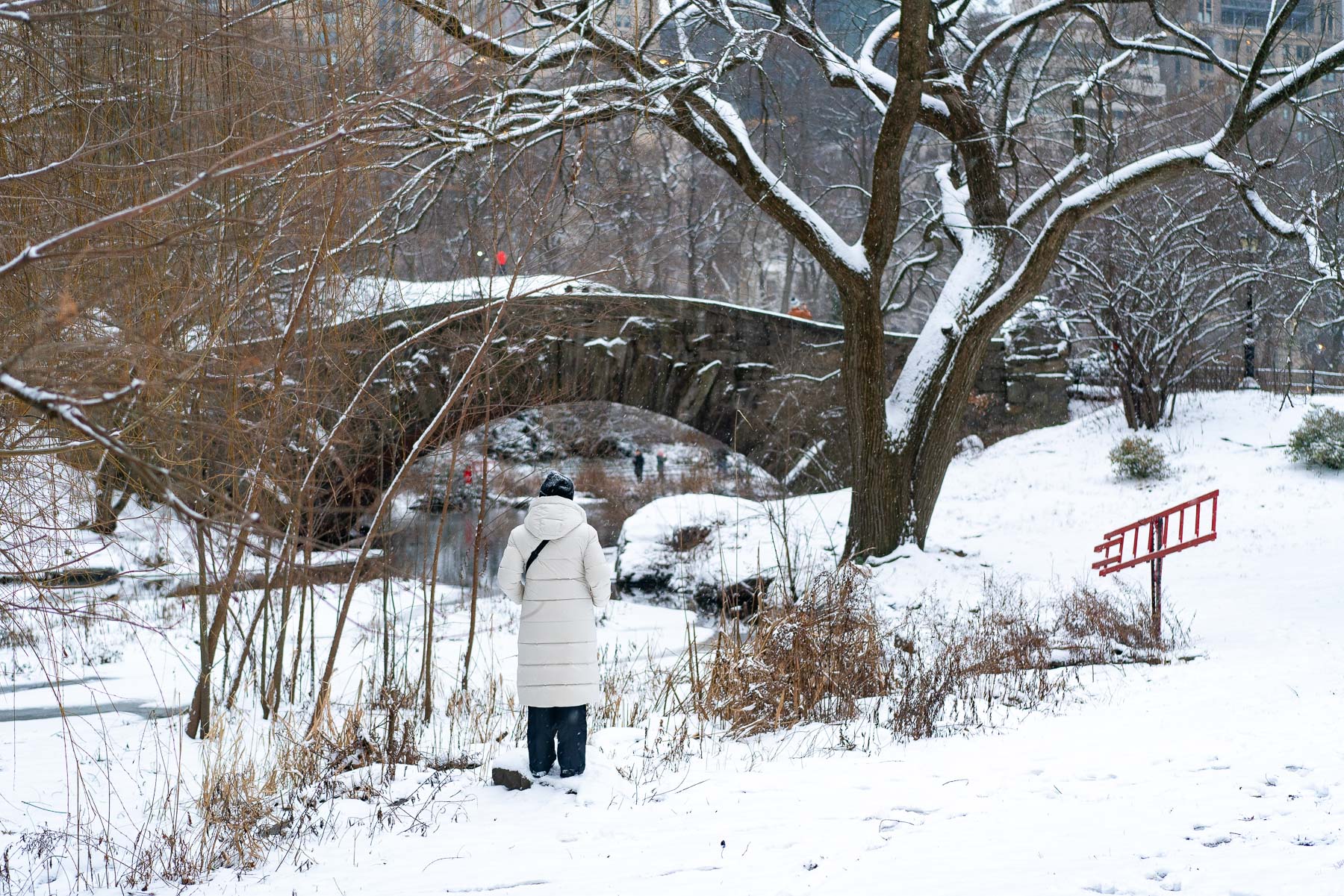 A woman in a white coat stands near the snow-covered Gapstow Bridge in Central Park.