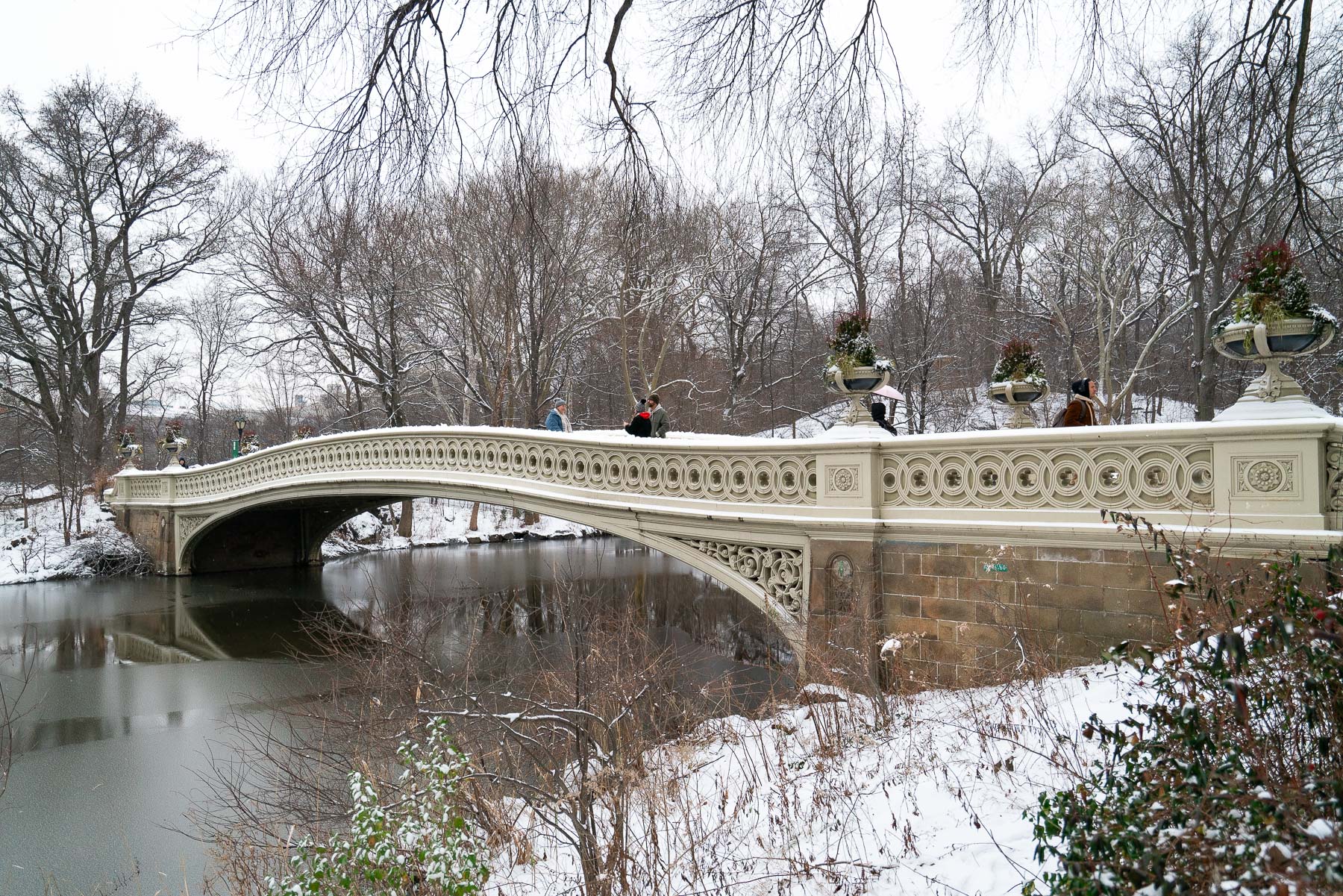Snow covers the ground around Bow Bridge in Central Park as a few people cross it.