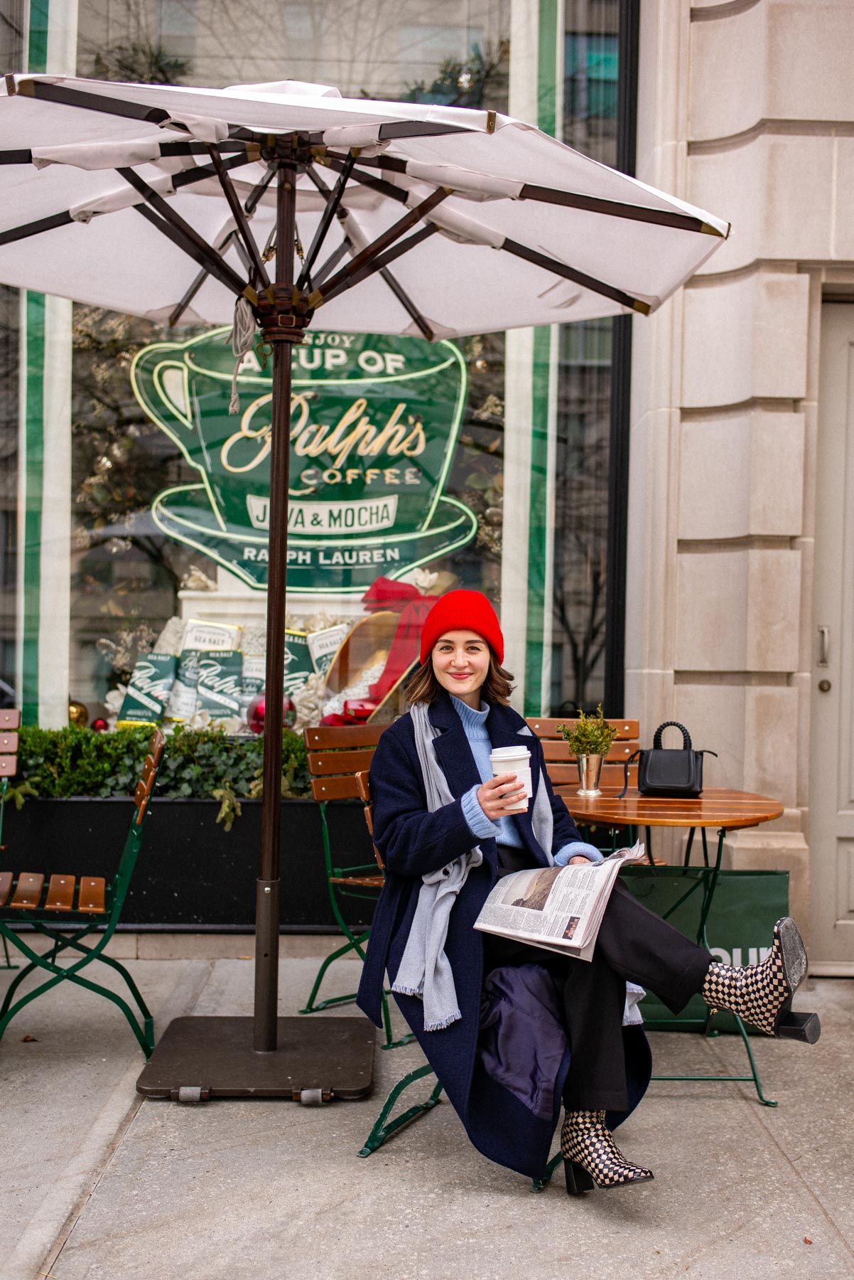 A woman sits at an outdoor table under an umbrella outside Ralph's Coffee, holding a cup and a newspaper.