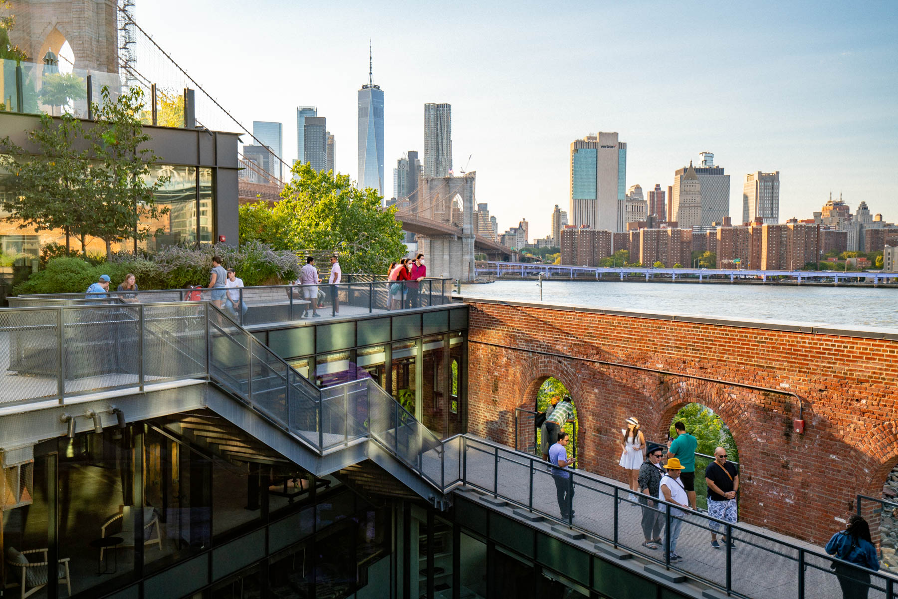 Visitors walk along an elevated rooftop terrace with brick archways overlooking the Manhattan skyline and Brooklyn Bridge across the East River.