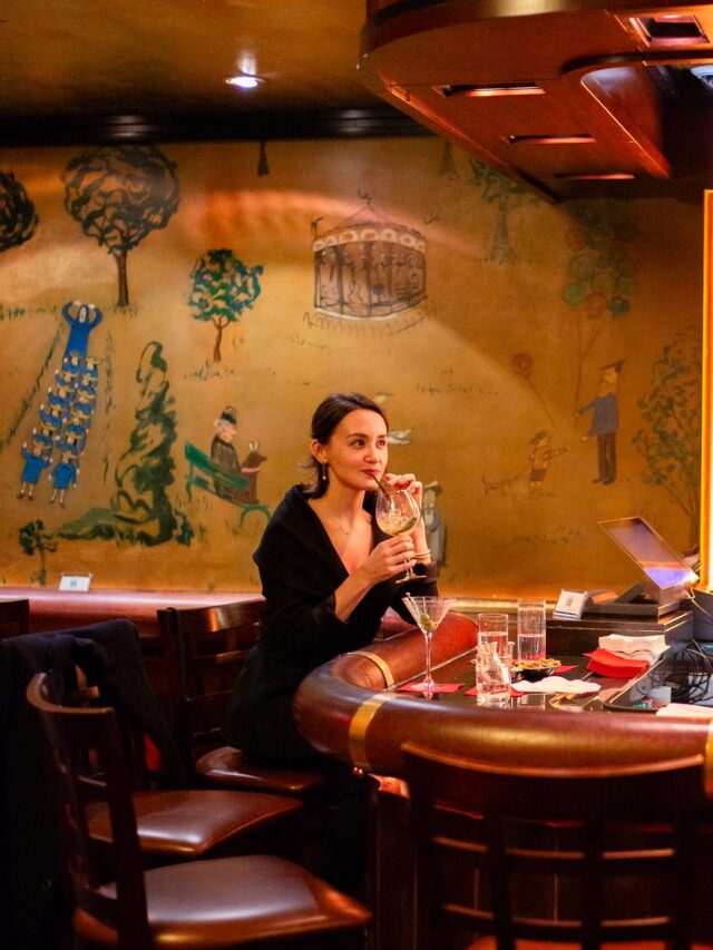 A woman sips a cocktail at the bar of Bemelmans Bar beneath its whimsical Central Park murals.