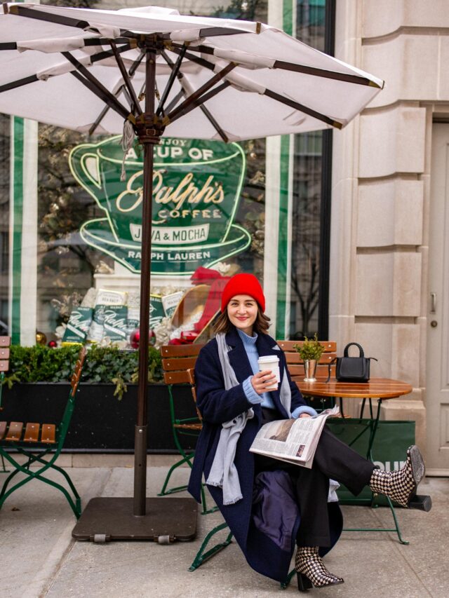 A woman sits at an outdoor table under an umbrella outside Ralph's Coffee holding a cup and a newspaper.