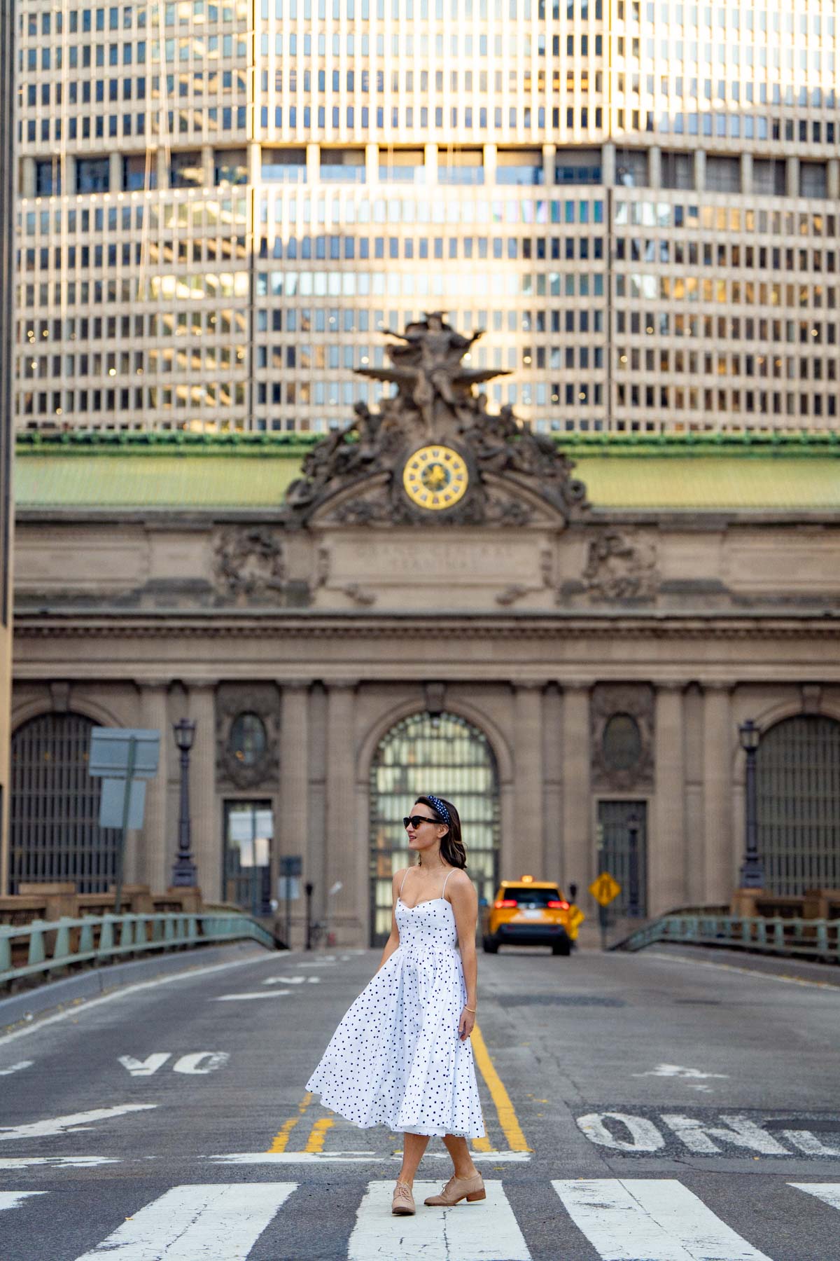 A woman in a polka dot dress stands on Park Avenue Viaduct in front of Grand Central Terminal's façade.