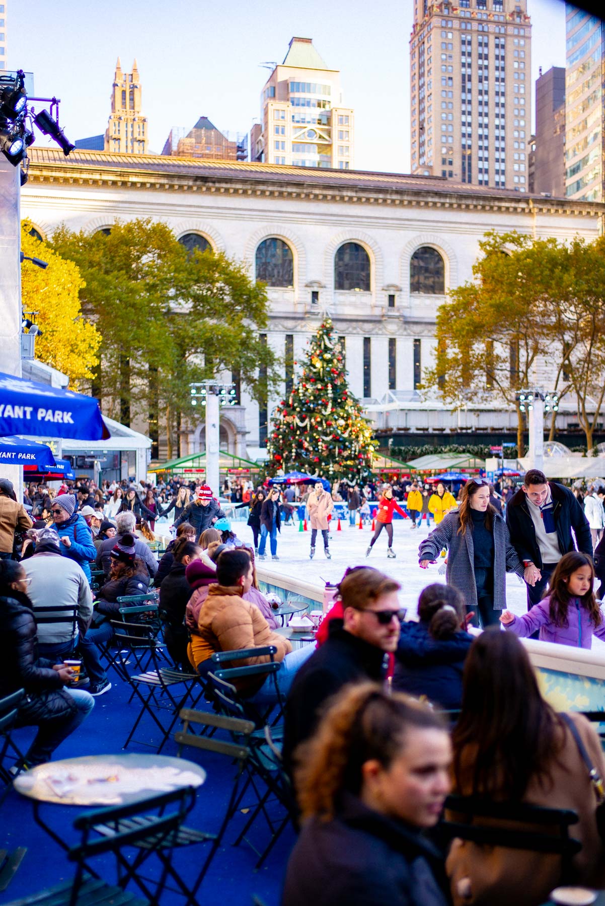 People skate and gather around a Christmas tree at an ice rink in Bryant Park with the New York Public Library in the background.