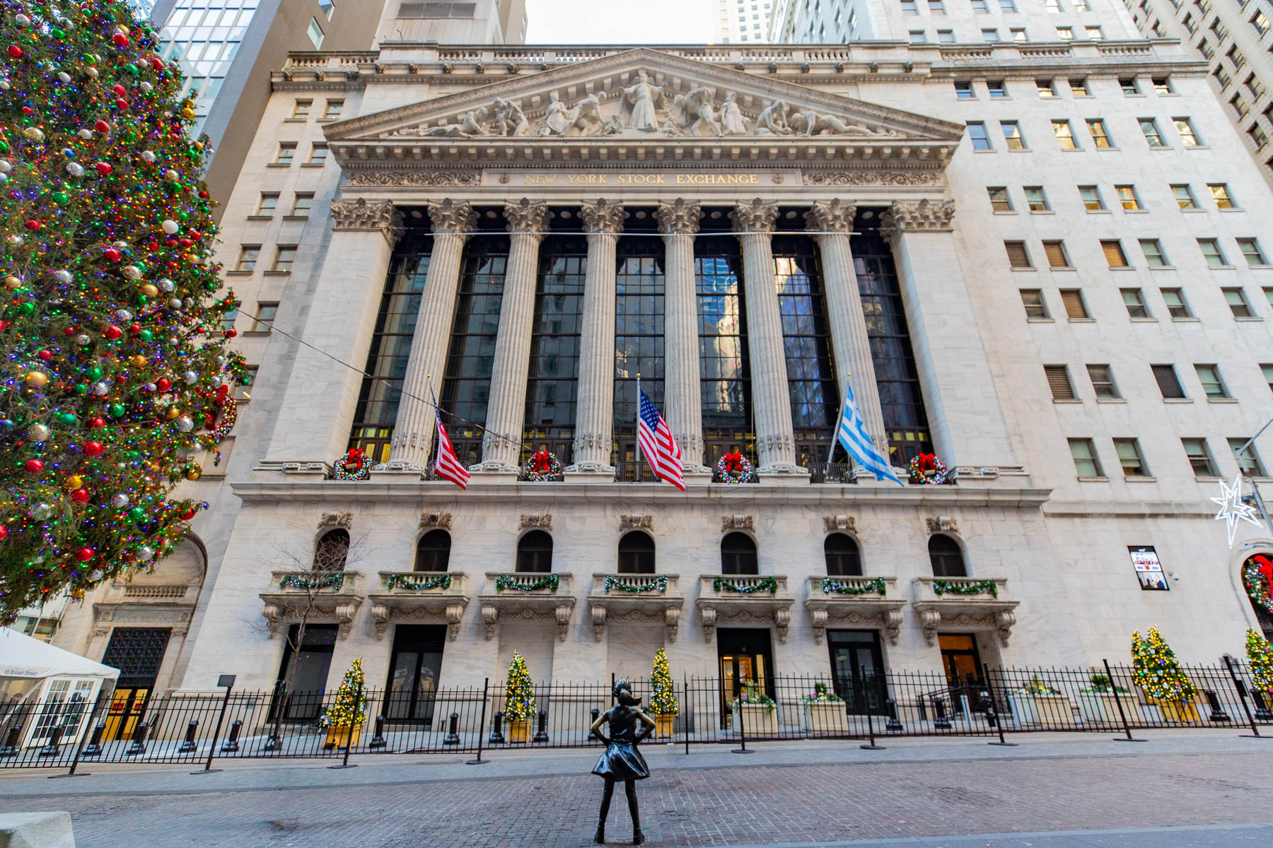 The New York Stock Exchange building is decorated with a large Christmas tree and wreaths, with the Fearless Girl statue standing in front.