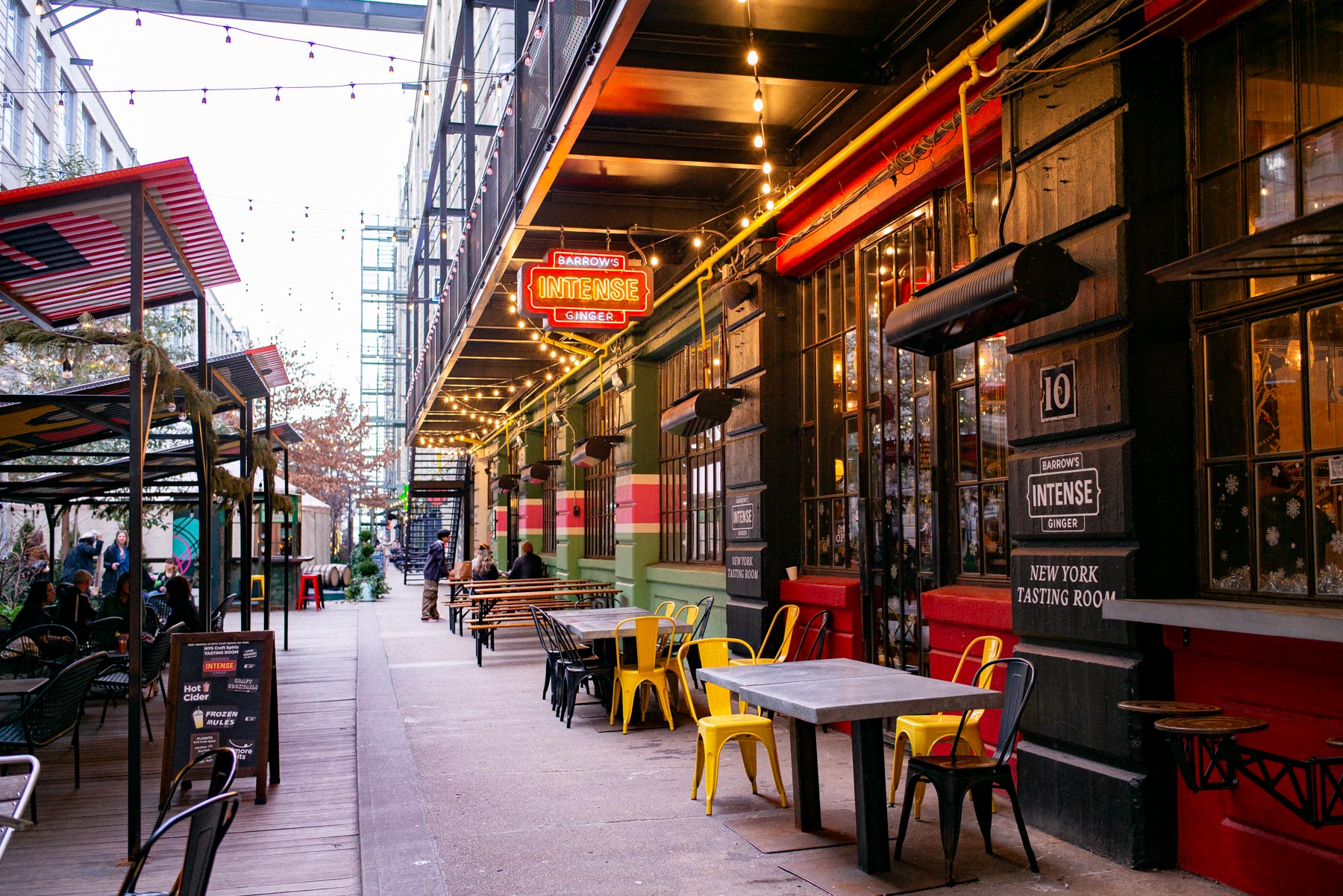 An outdoor seating area lines a covered walkway at Industry City with a neon sign reading Barrow's Intense Ginger.