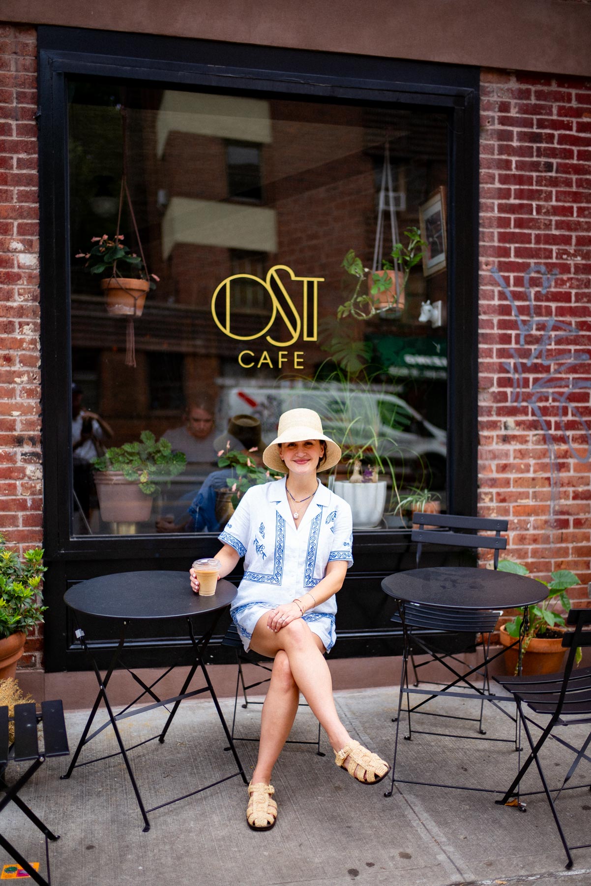 A woman sits at an outdoor table in front of Ost Cafe holding an iced coffee.