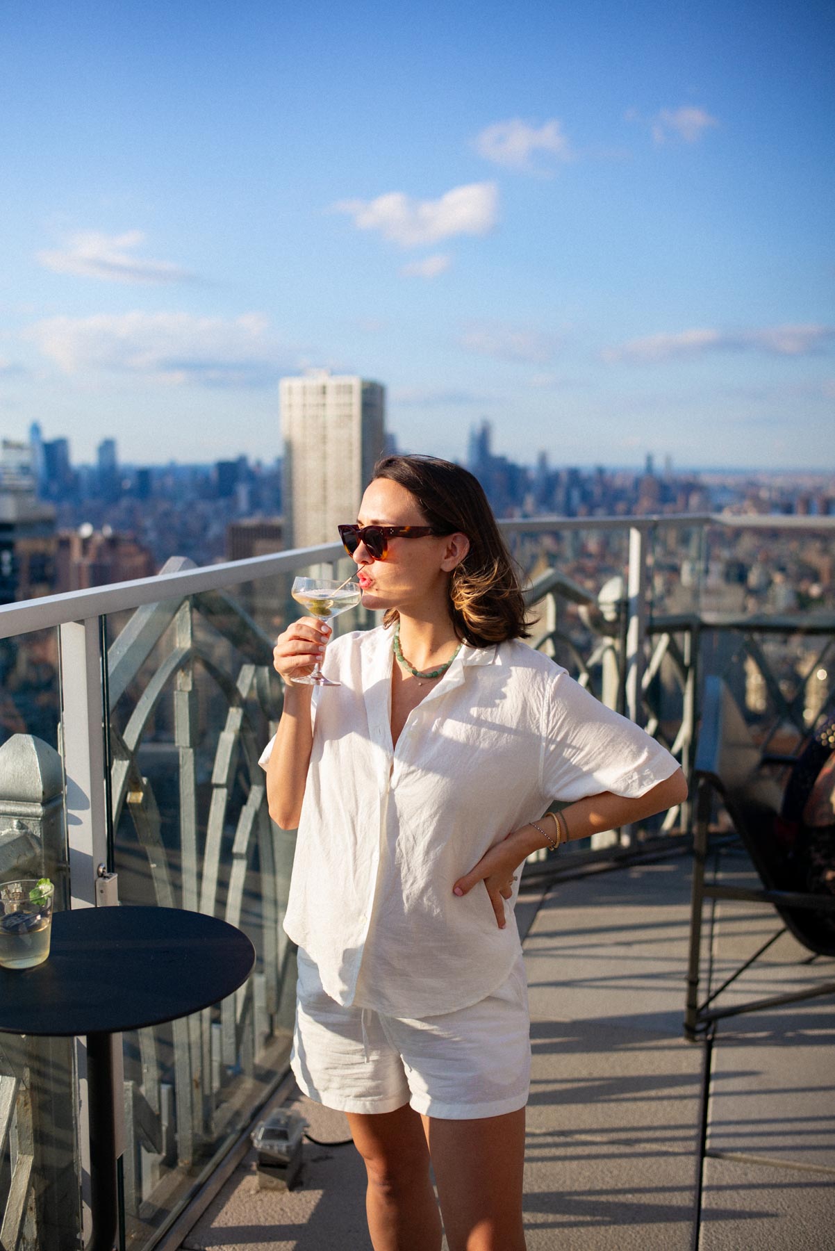 A woman in white sips a cocktail on a rooftop bar overlooking the city skyline.