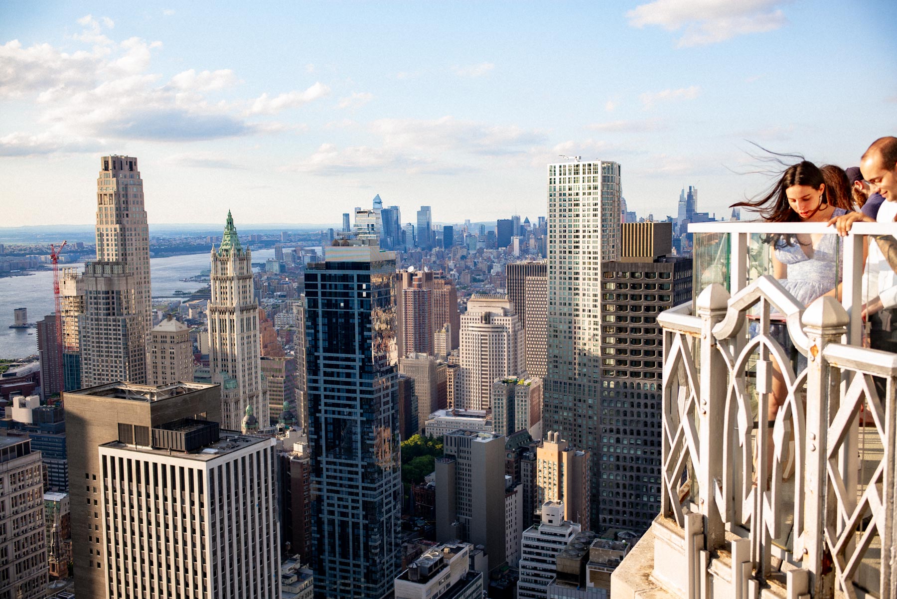 People lean on a rooftop bar railing overlooking the lower Manhattan skyline.