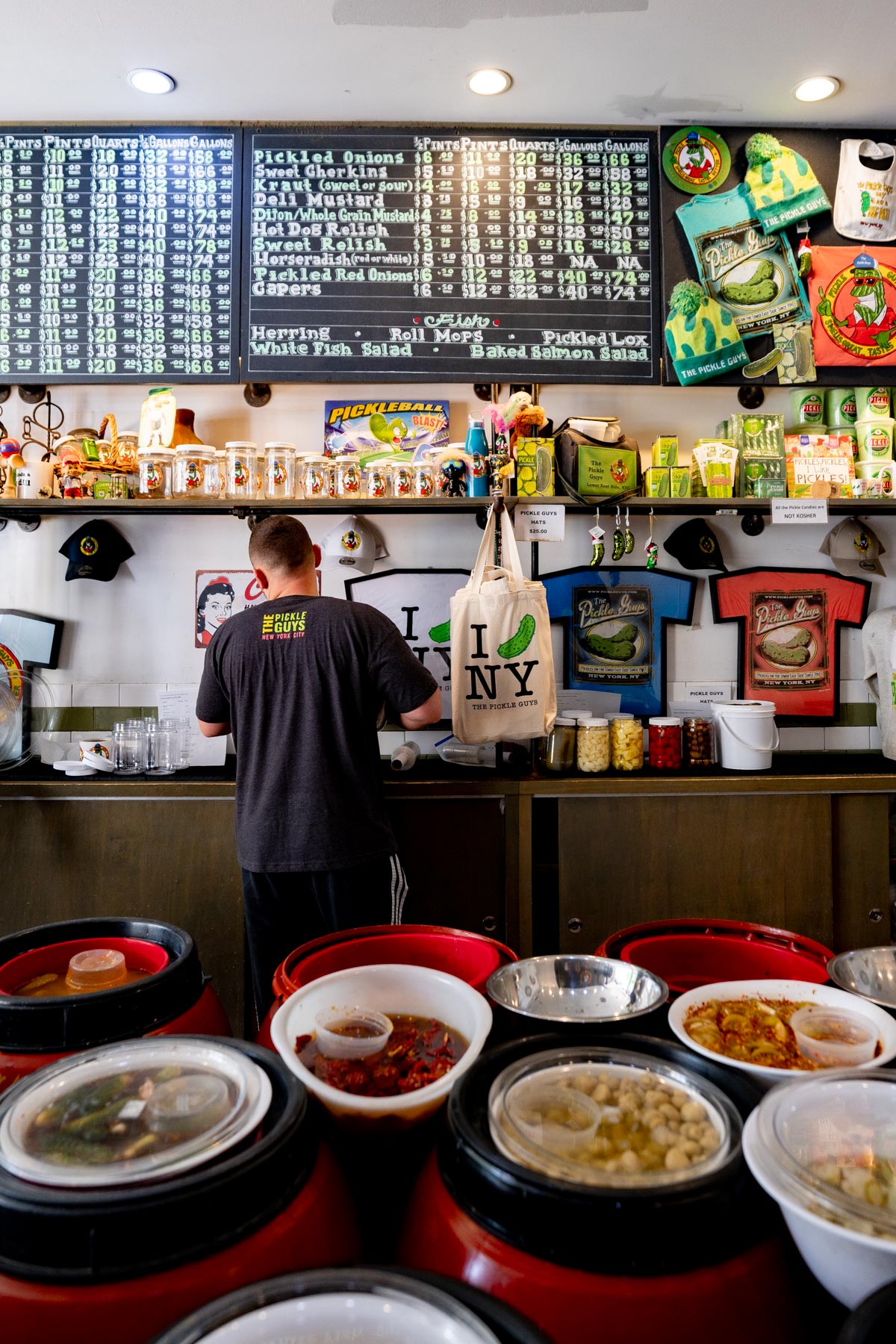 A worker stands behind the counter at The Pickle Guys shop surrounded by barrels of pickles and shelves of merchandise.