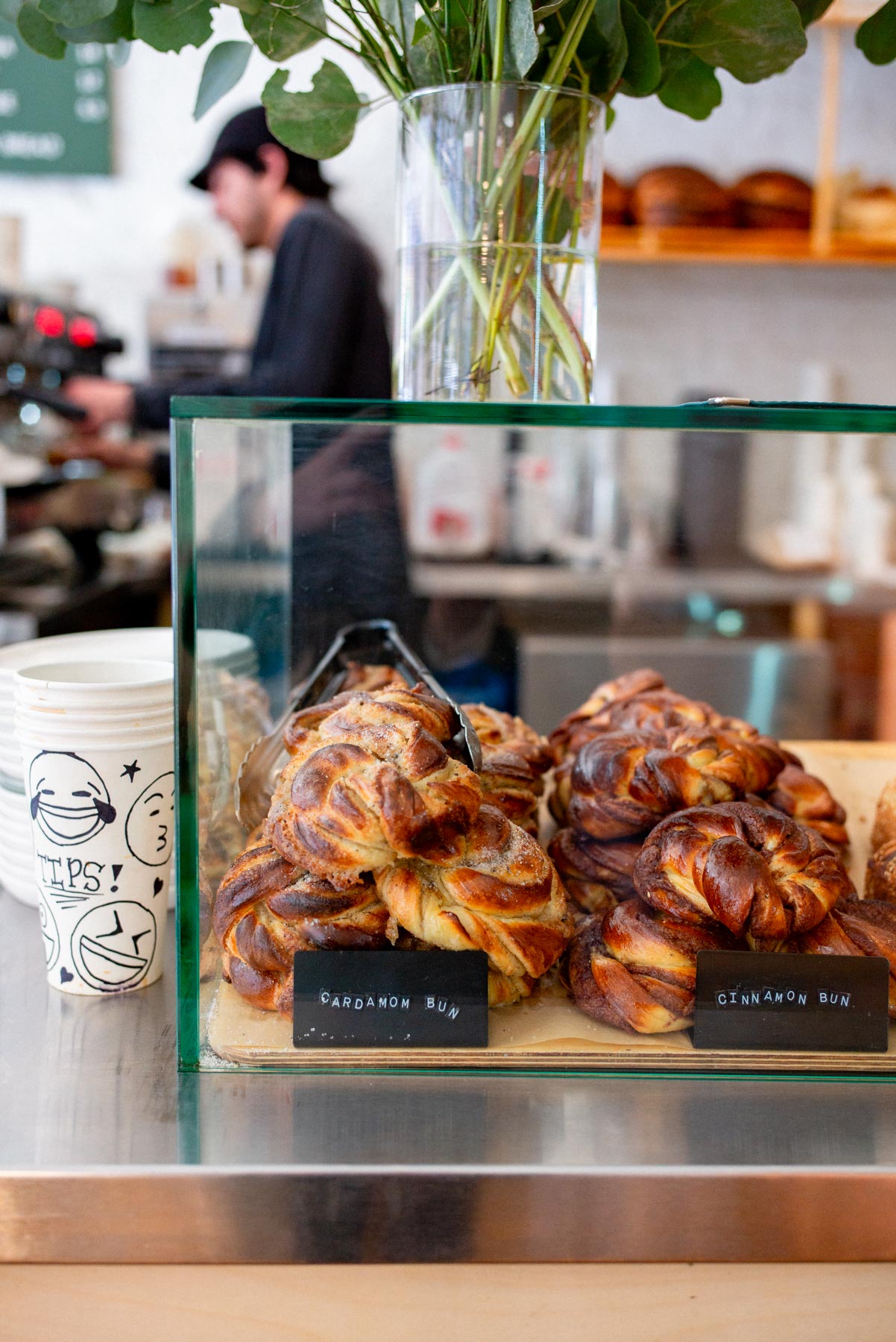 A glass display case holds cardamom and cinnamon buns at a café counter with a barista working behind it.