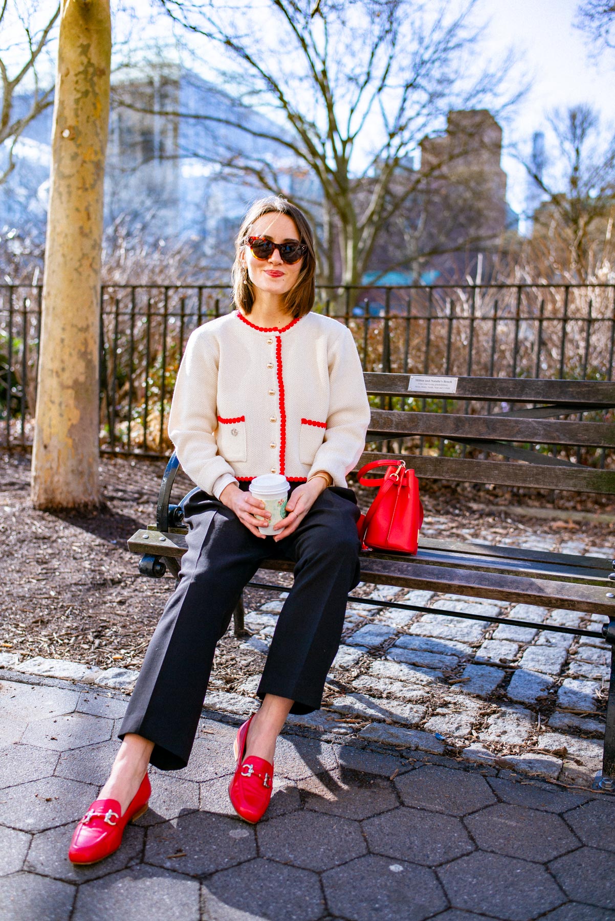 Trendy fashion woman sitting on a park bench with a bright red purse, red loafers, and red detailed cardigan 