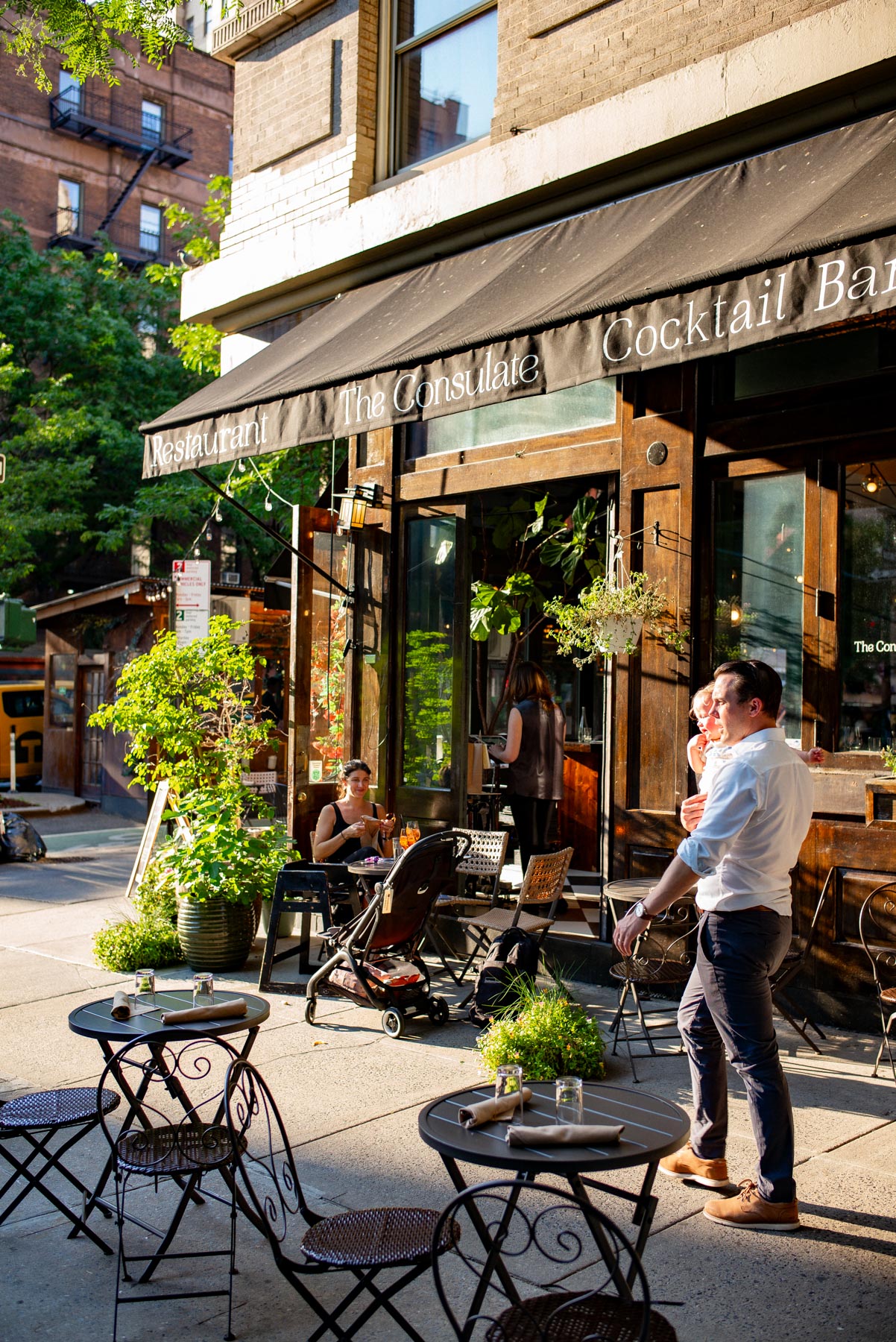A restaurant storefront called The Consulate has outdoor cafe tables on the sidewalk with people seated and standing nearby.
