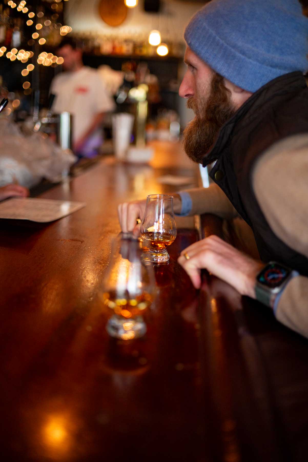 A bearded man in a beanie sits at a bar with a whisky flight in front of him.