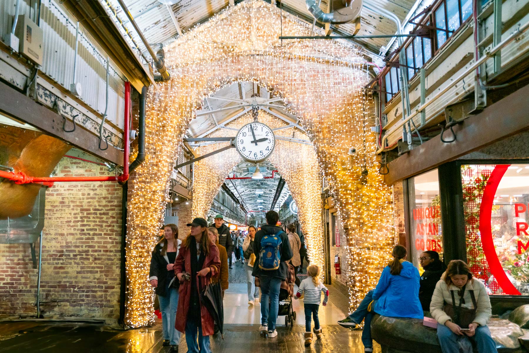 People walk through the string-light-covered corridor of Chelsea Market beneath a large clock.