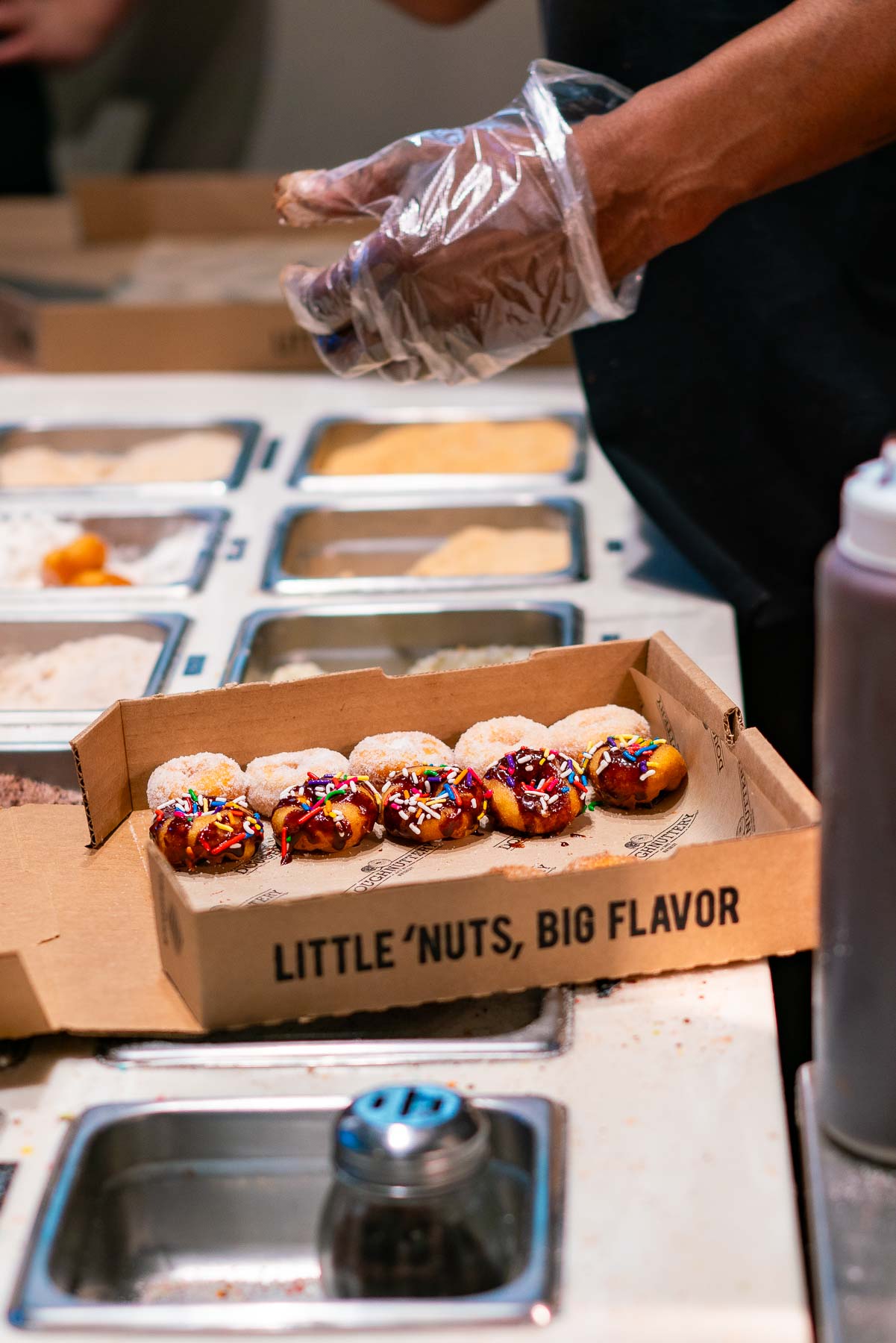 A worker sprinkles toppings on a box of mini doughnuts at a market stall.
