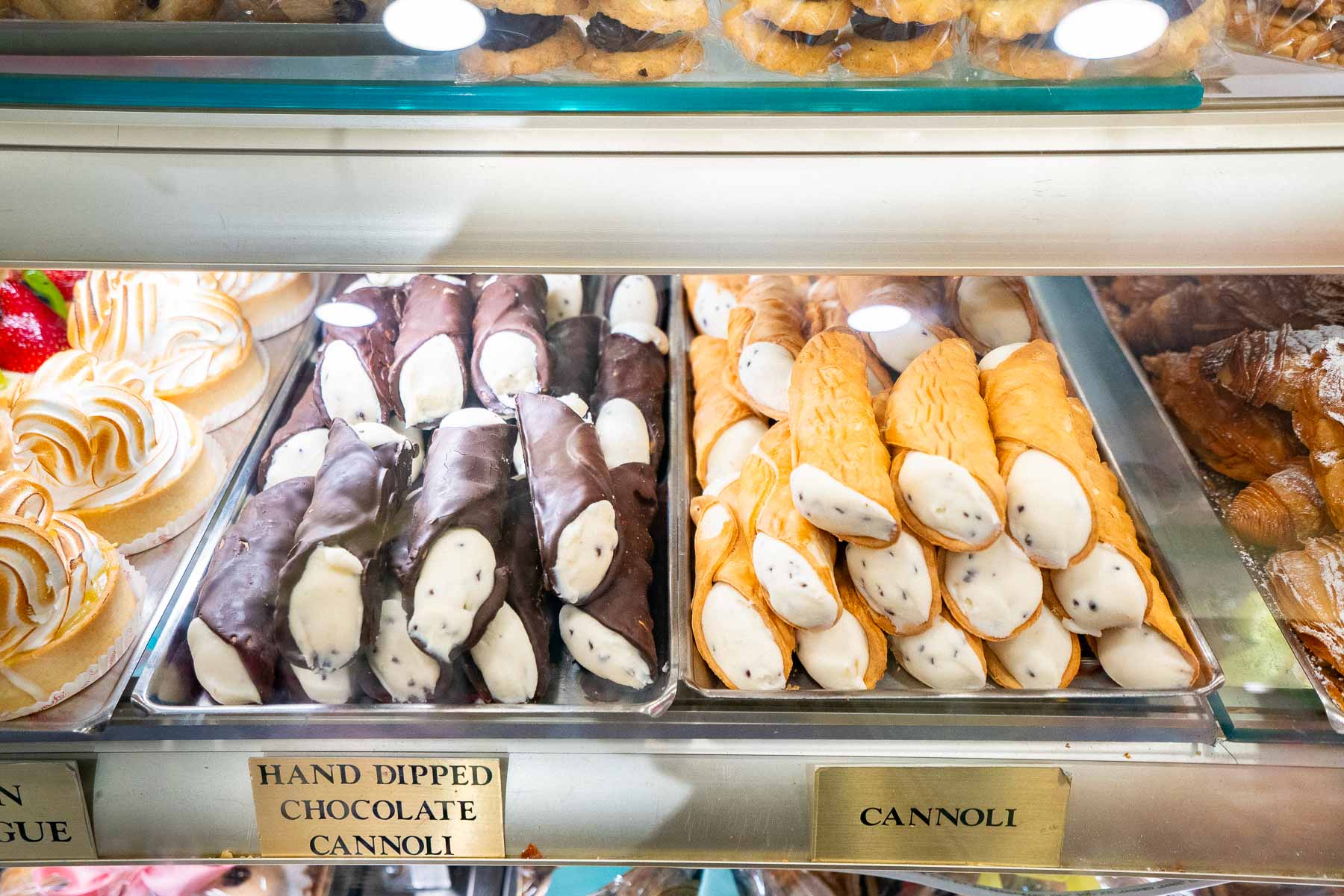 A pastry case displays cannoli, chocolate-dipped cannoli, meringues, and croissants at a bakery.