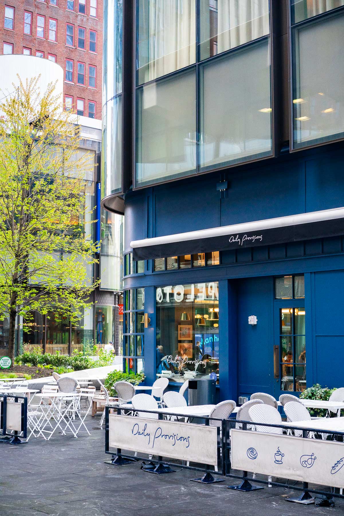 A blue-paneled storefront of Daily Provisions with outdoor cafe seating sits among glass towers.