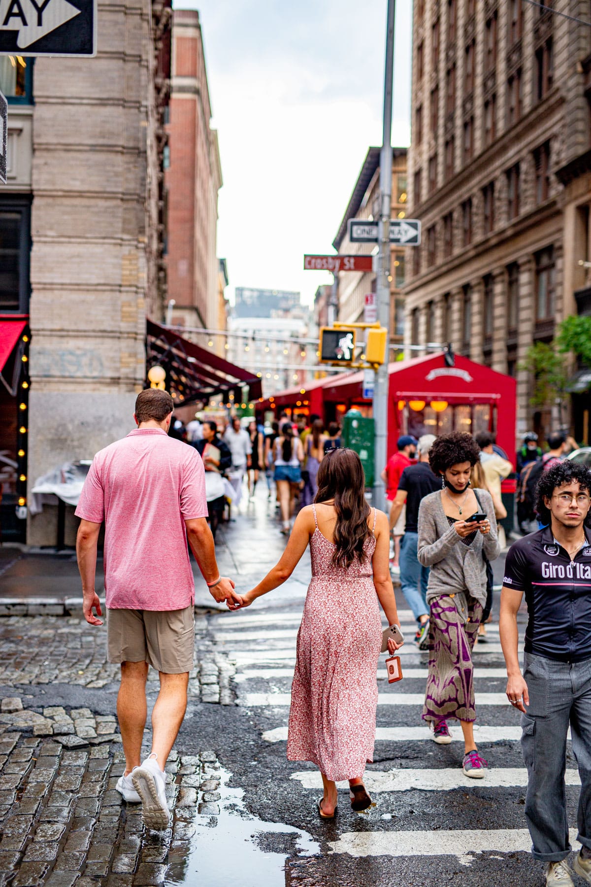 Couple wearing pink holding hands crossing a rainy crosswalk in SoHo heading towards the red canopy at Balthazars in New York City